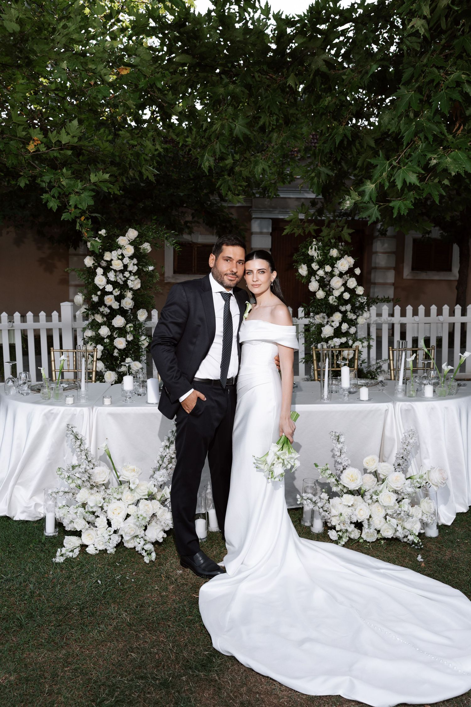 Elegant outdoor wedding ceremony setup with white floral arches, picket fence, and decorated tables in a garden setting.