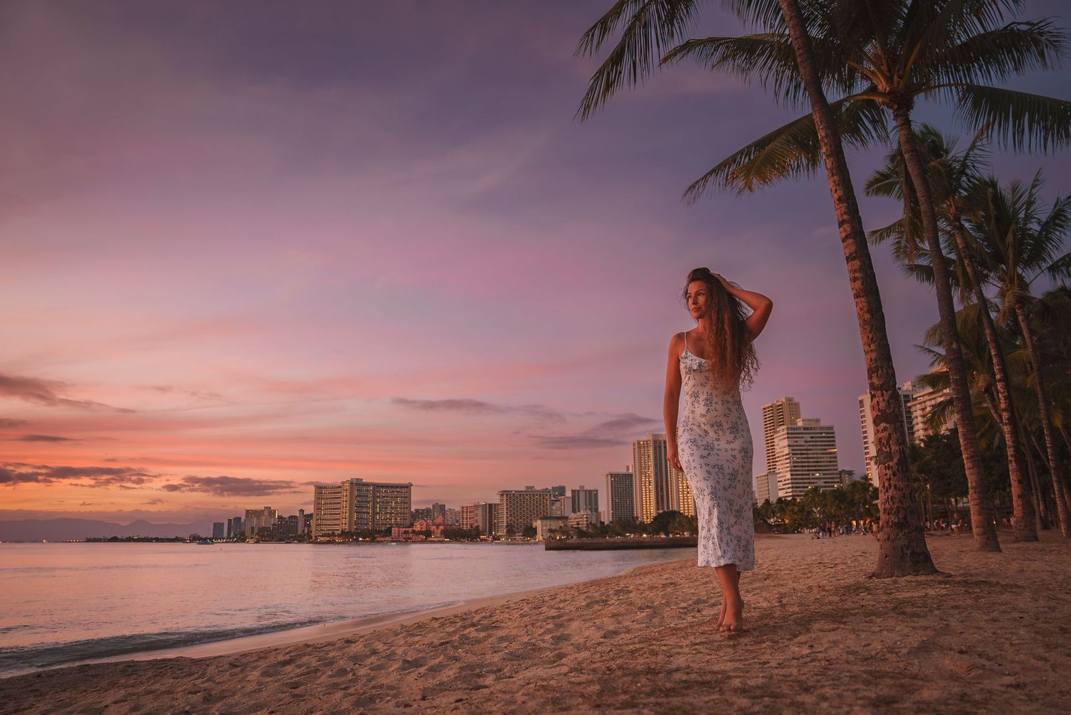 Tropical beach panorama with palm trees and city skyline silhouetted against a vibrant sunset sky.