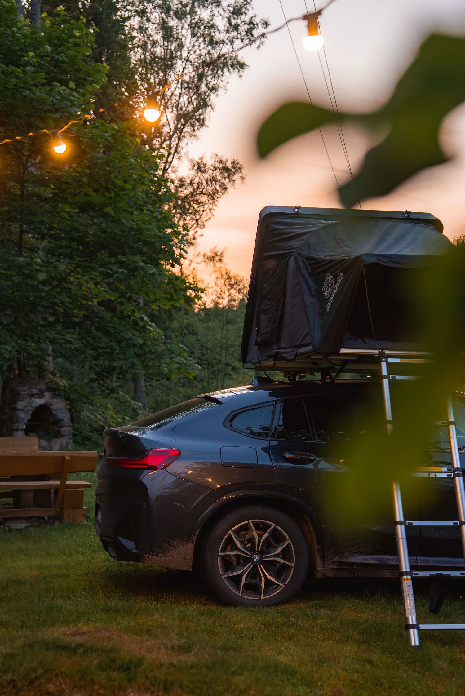 Rooftop tent camping setup with string lights at dusk in wooded area.