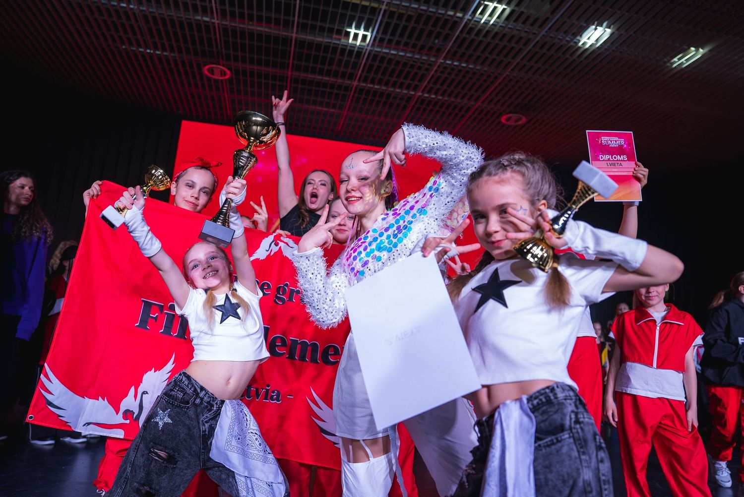 Dancers perform energetic choreography in white costumes against a red-lit stage backdrop.