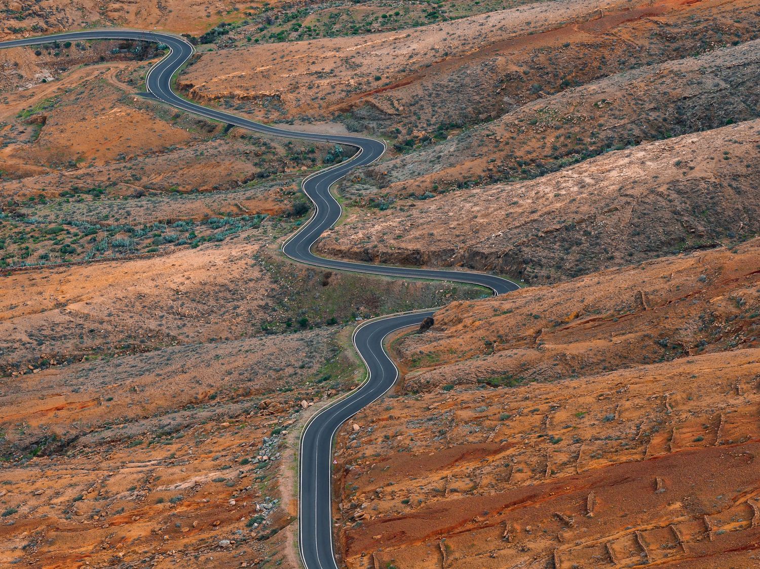 A winding road curves through red desert landscape with steep cliffs and rugged terrain in the American Southwest.