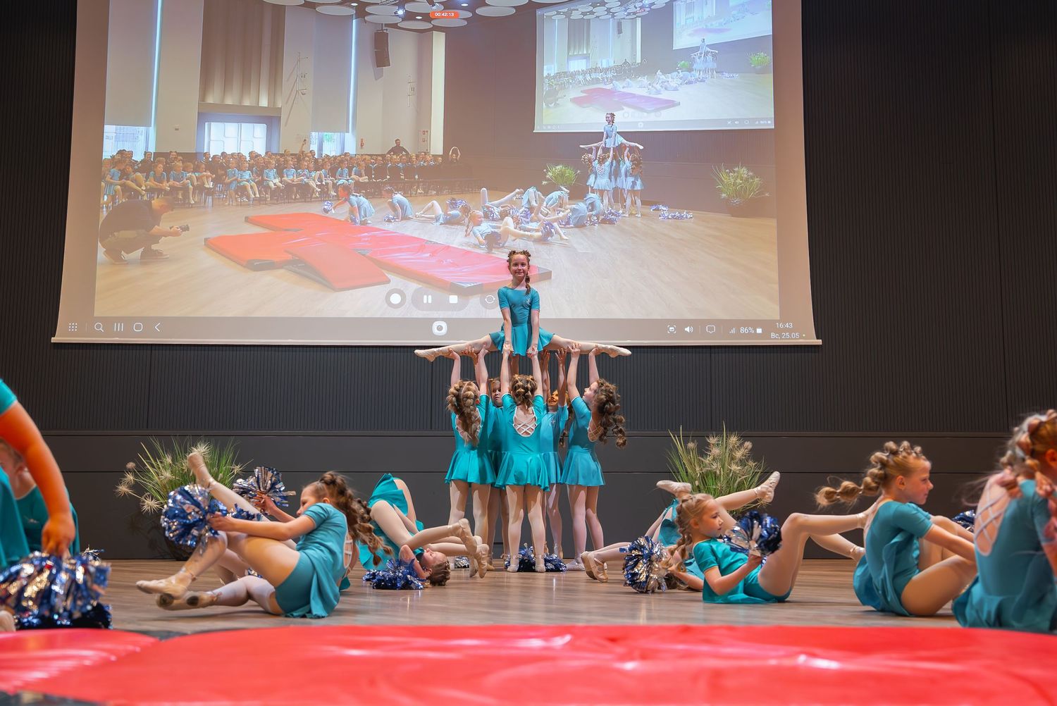 Dance performance with multiple performers in matching turquoise costumes on a dark stage.