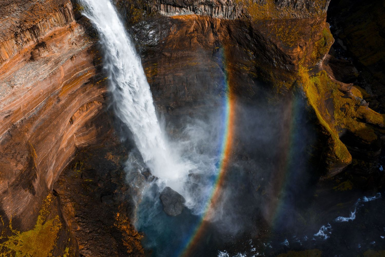 A majestic waterfall cascades down orange canyon walls creating a vibrant rainbow in the mist below.