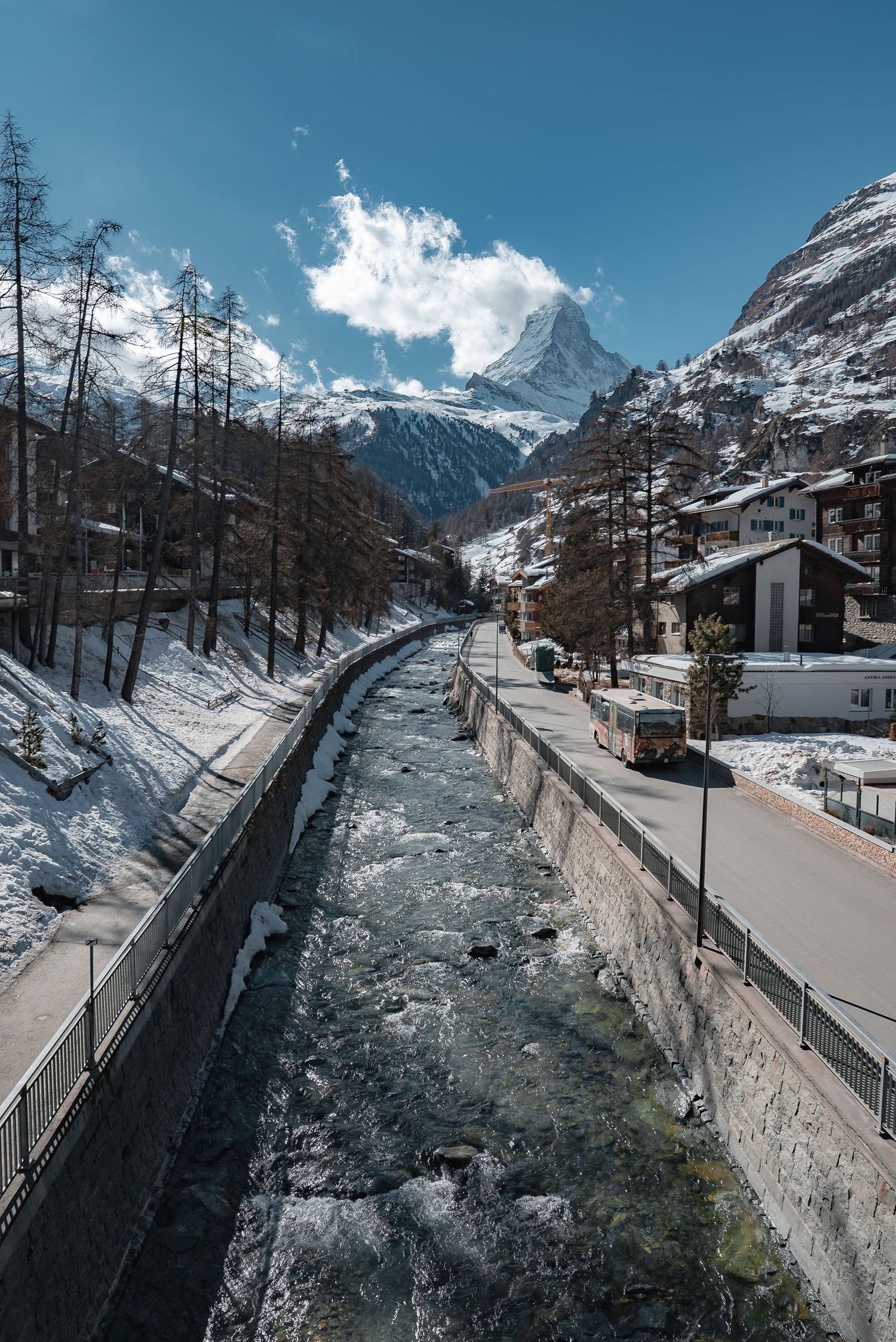 Snow-covered mountain road with dramatic alpine peaks and stone wall barrier in winter.