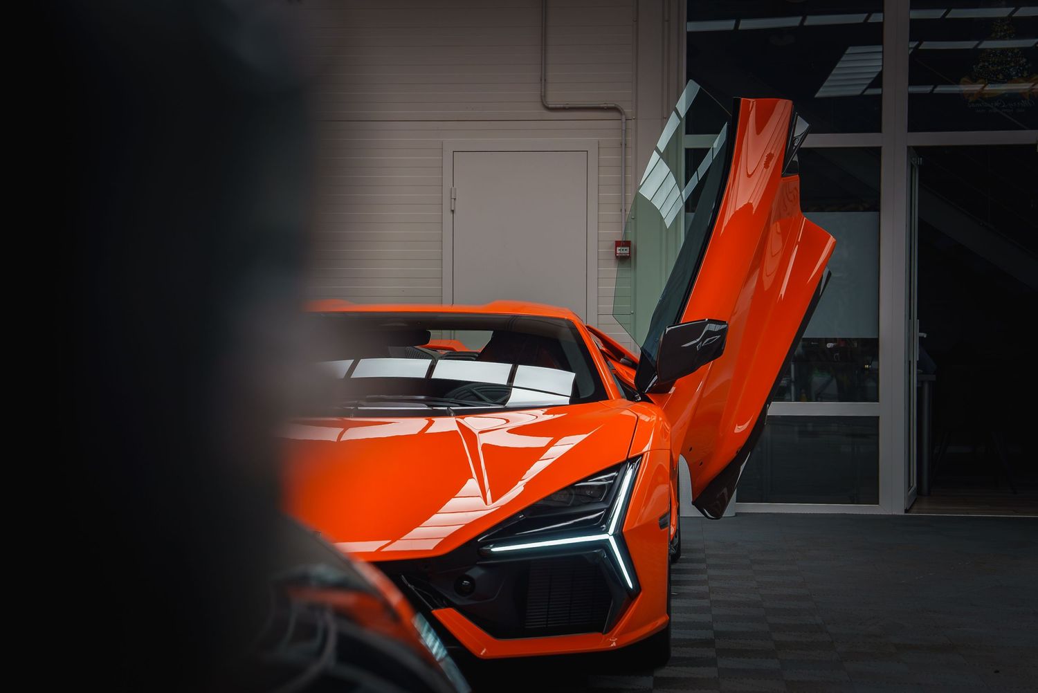 Orange Lamborghini sports car with dramatic upward-opening doors in dark garage.