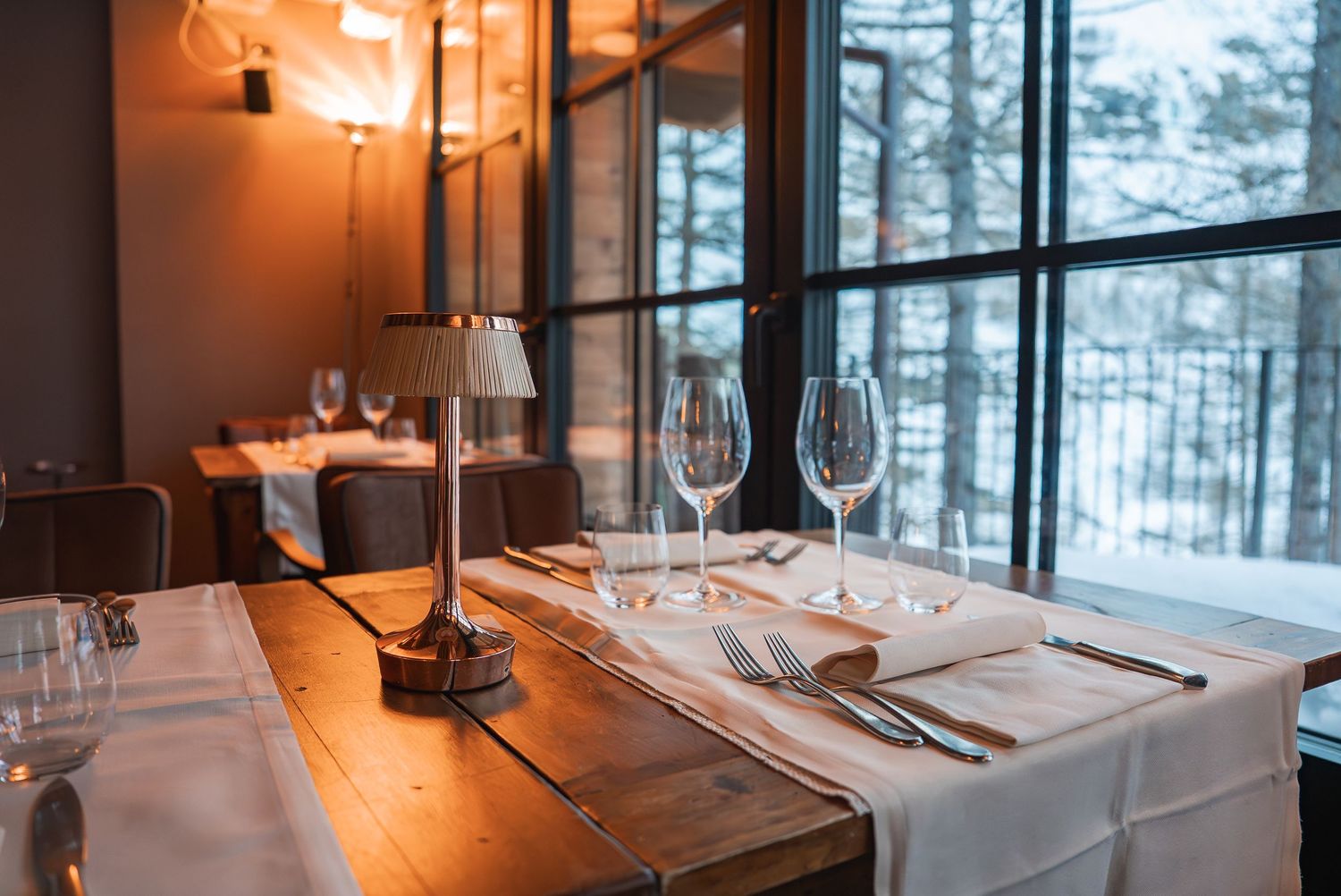 Intimate restaurant setting with warm lighting and large windows overlooking a snowy forest.