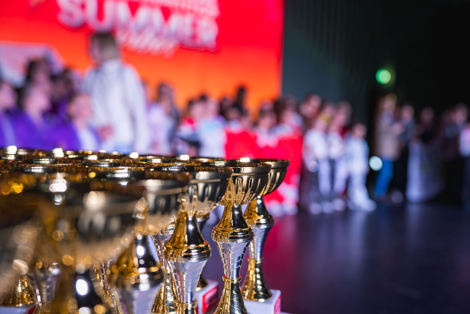 Row of golden trophies displayed at dance competition with red stage lighting in background.