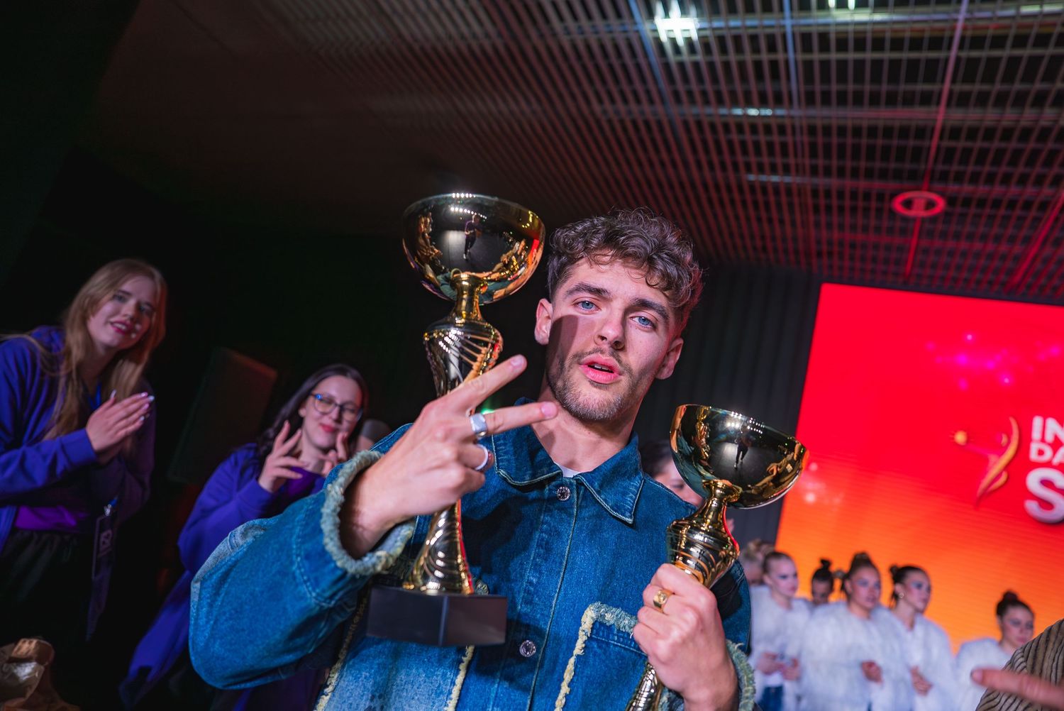 Performers celebrate on stage at an international dance competition with trophies and bright red lighting.