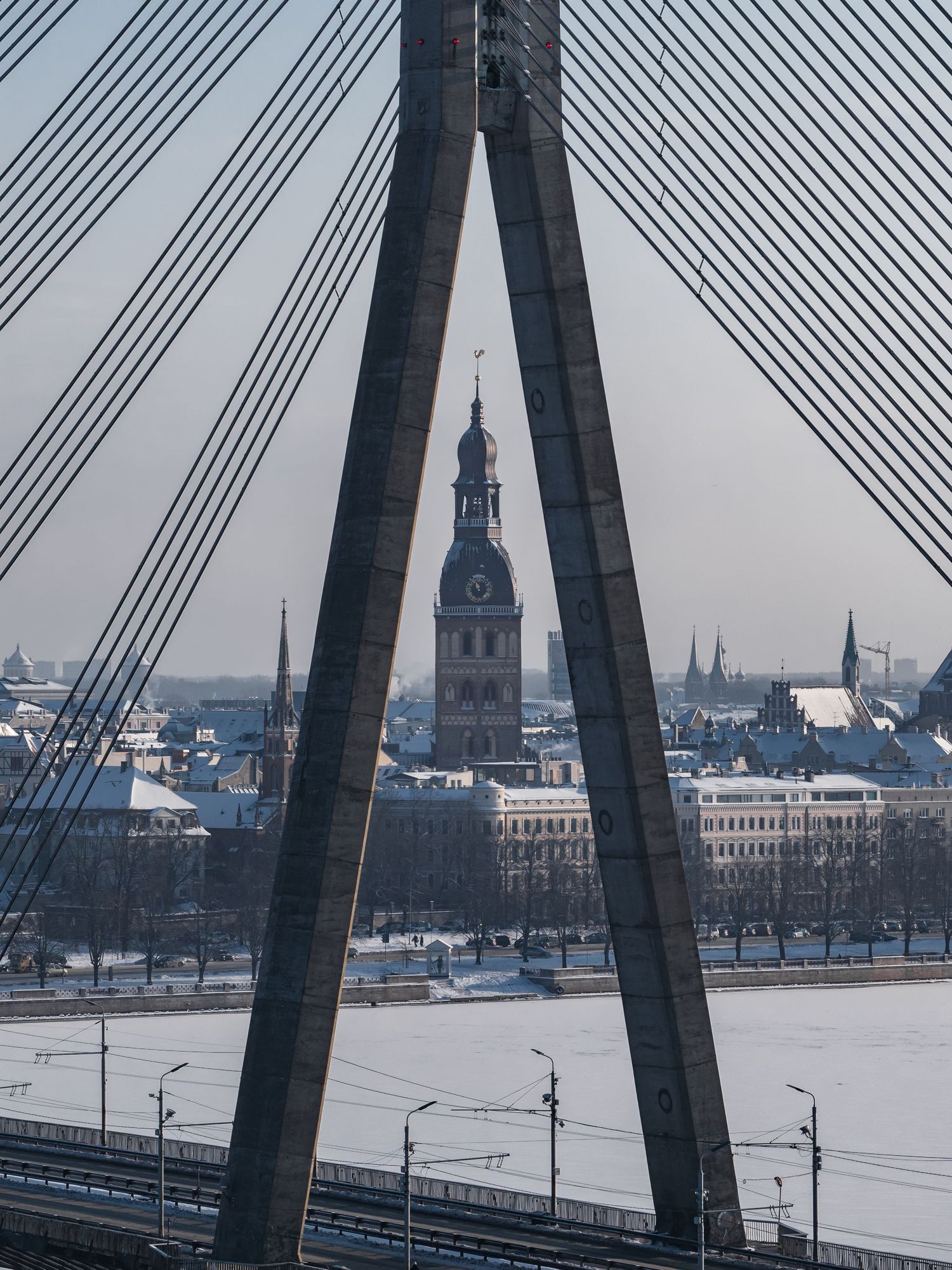 Winter cityscape of Riga, Latvia with snow-covered Daugava River and historic church spires visible through bridge supports.