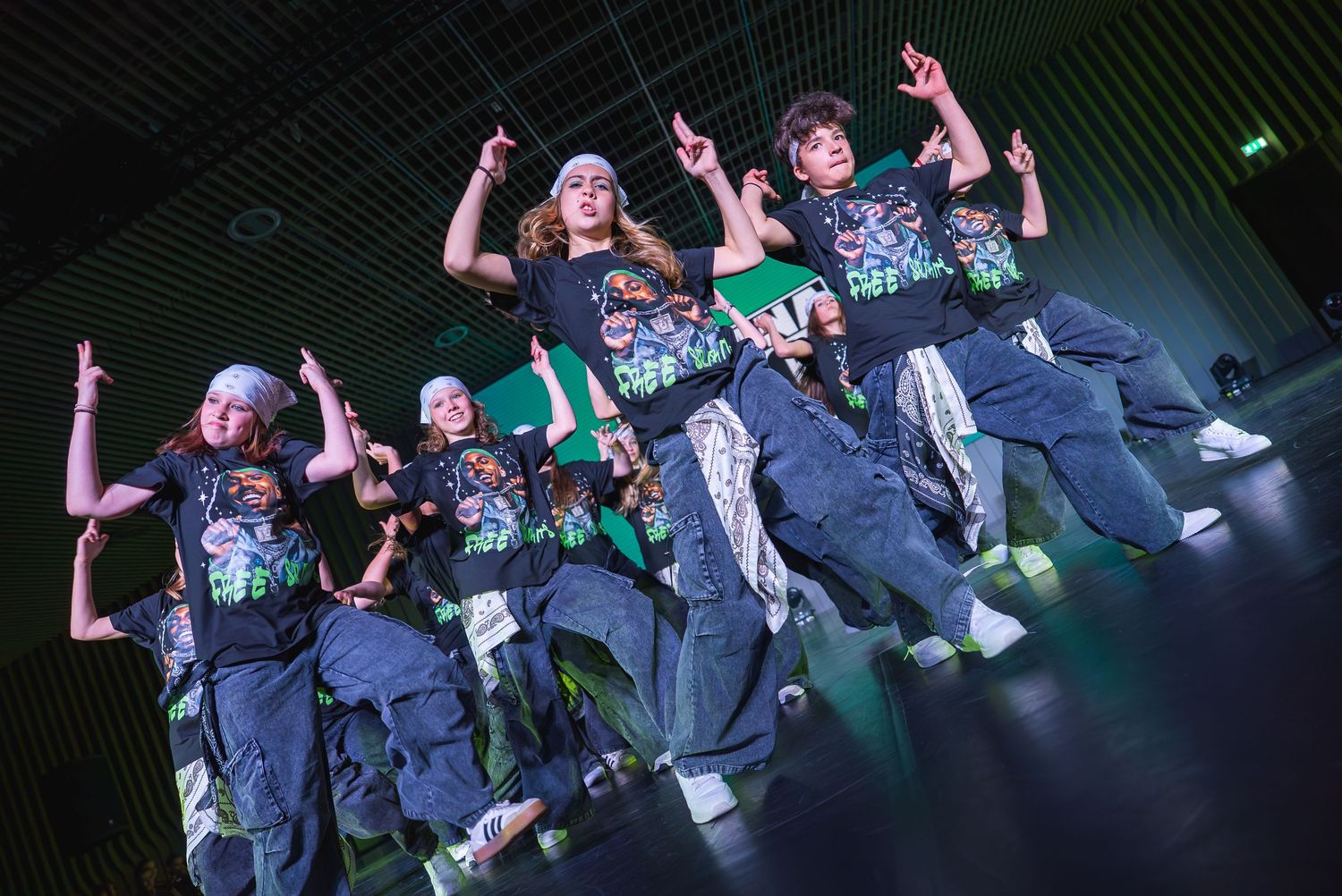 Group of performers executing synchronized dance moves in dark outfits under stage lights.