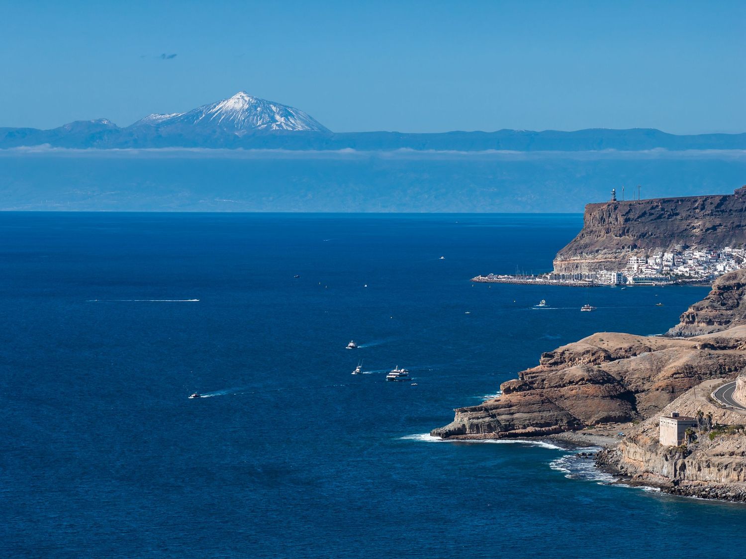 Scenic coastal view of rugged cliffs along deep blue waters with snow-capped Mount Baker visible in the distance.