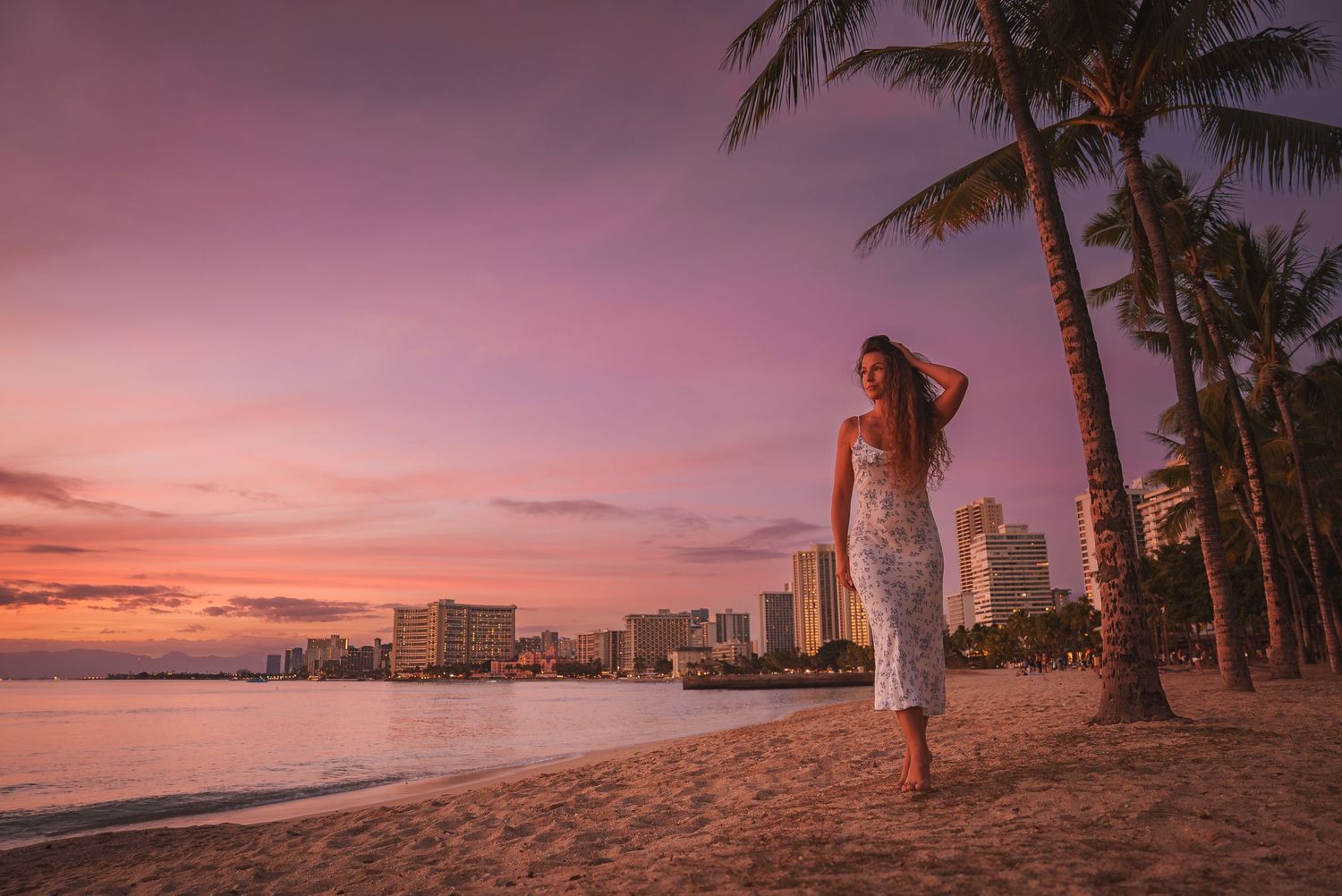 Wide-angle view of a Hawaiian beach at sunset with palm trees framing the coastal cityscape.