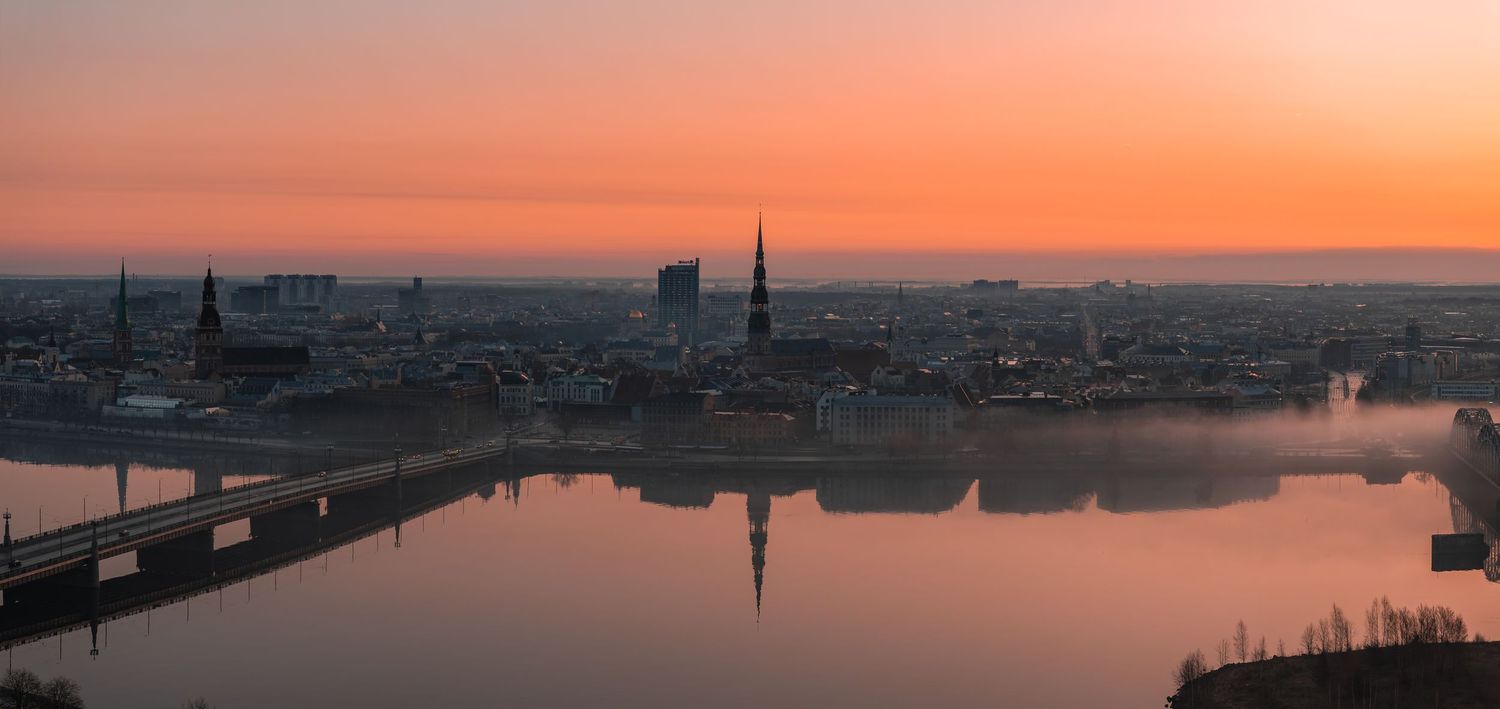Panoramic cityscape at sunset with church spires reflected in calm river waters.
