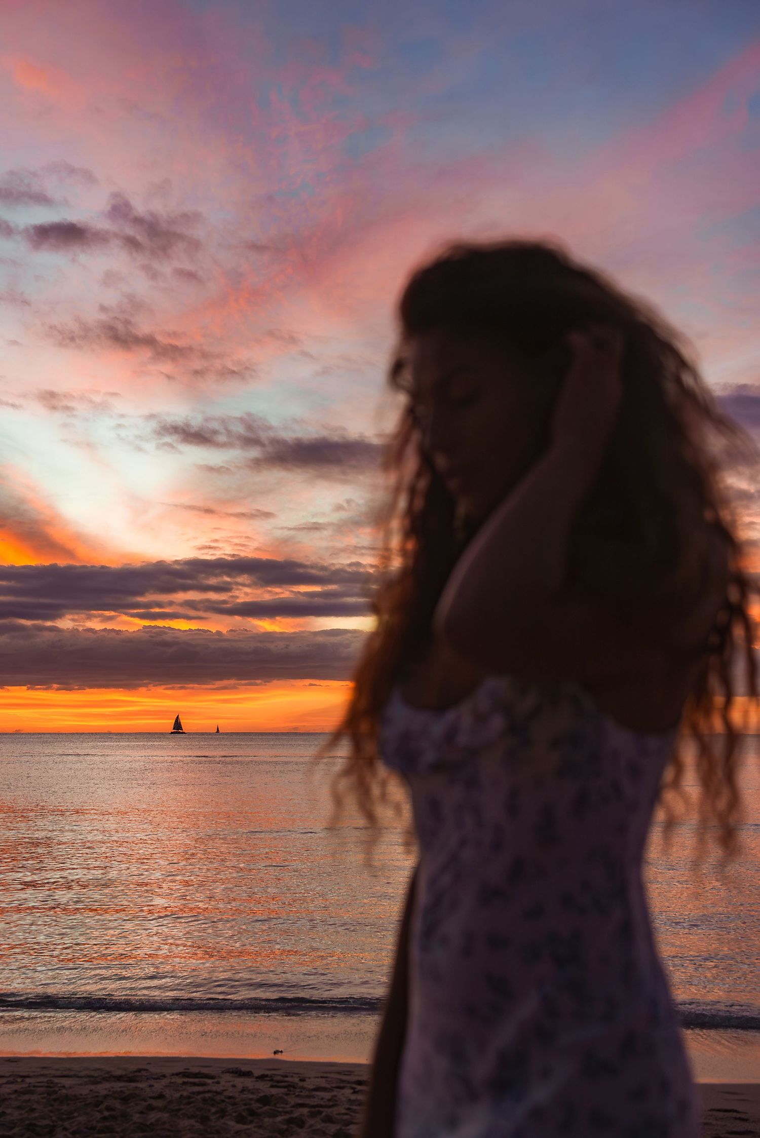 Silhouette against vibrant orange and pink sunset sky over ocean with sailboats on horizon.