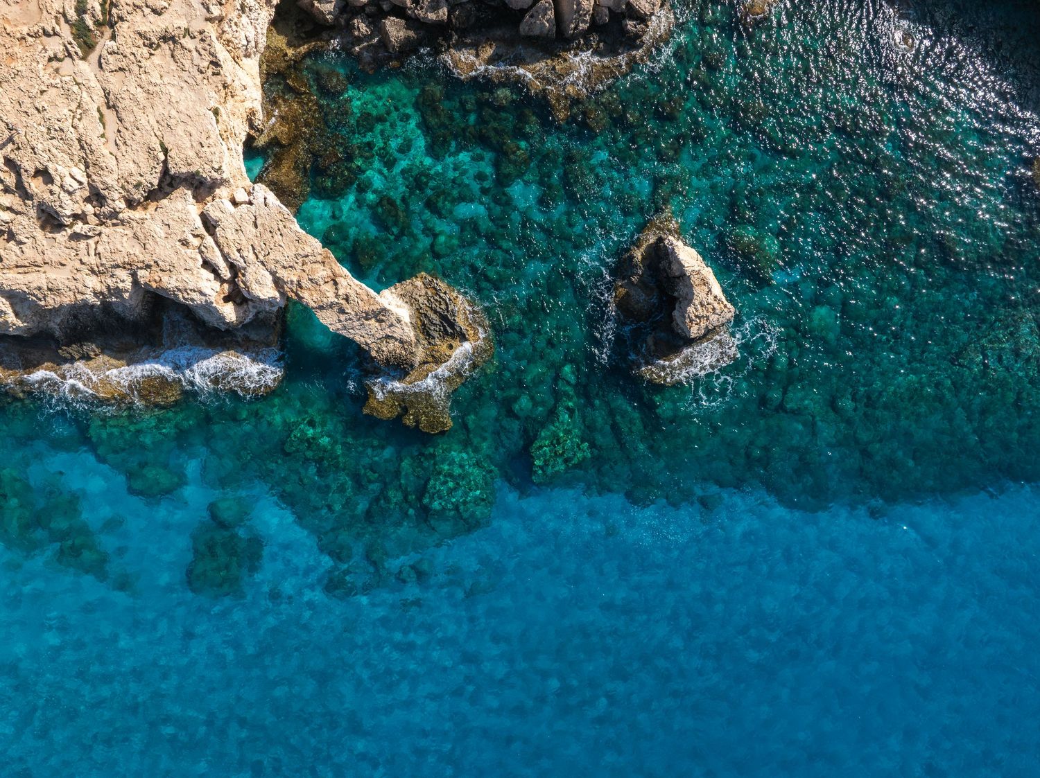 Aerial view of rocky coastline with crystal clear turquoise Mediterranean waters and rugged cliffs.