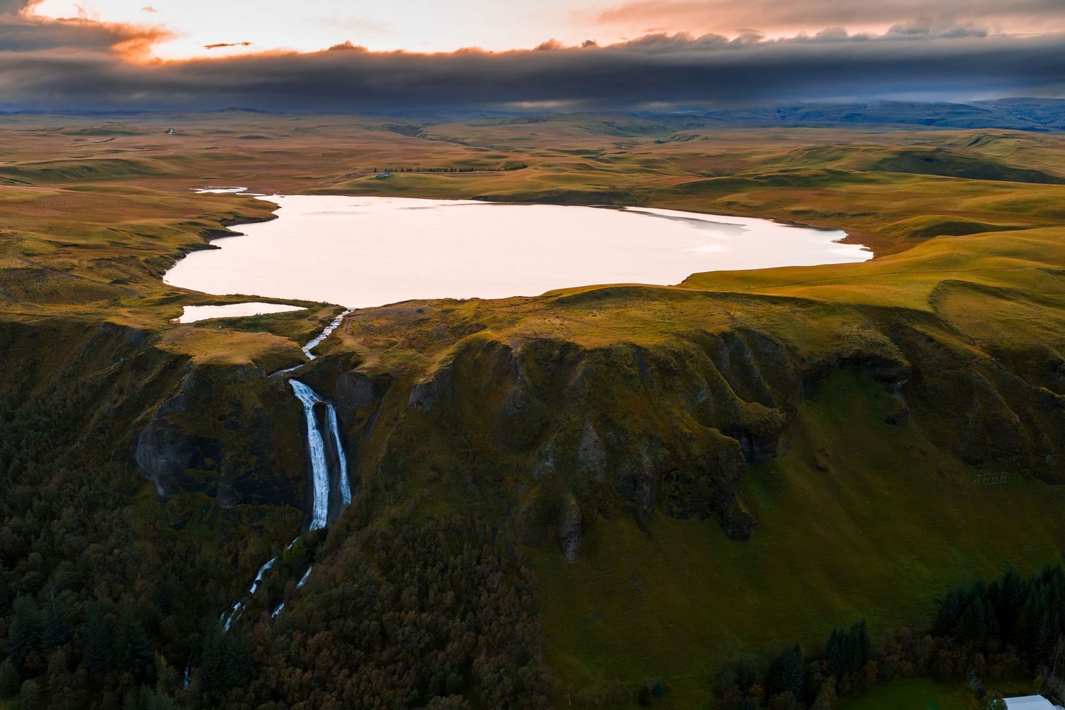 Aerial view of a dramatic waterfall flowing into a snow-covered lake surrounded by green hills during sunset.