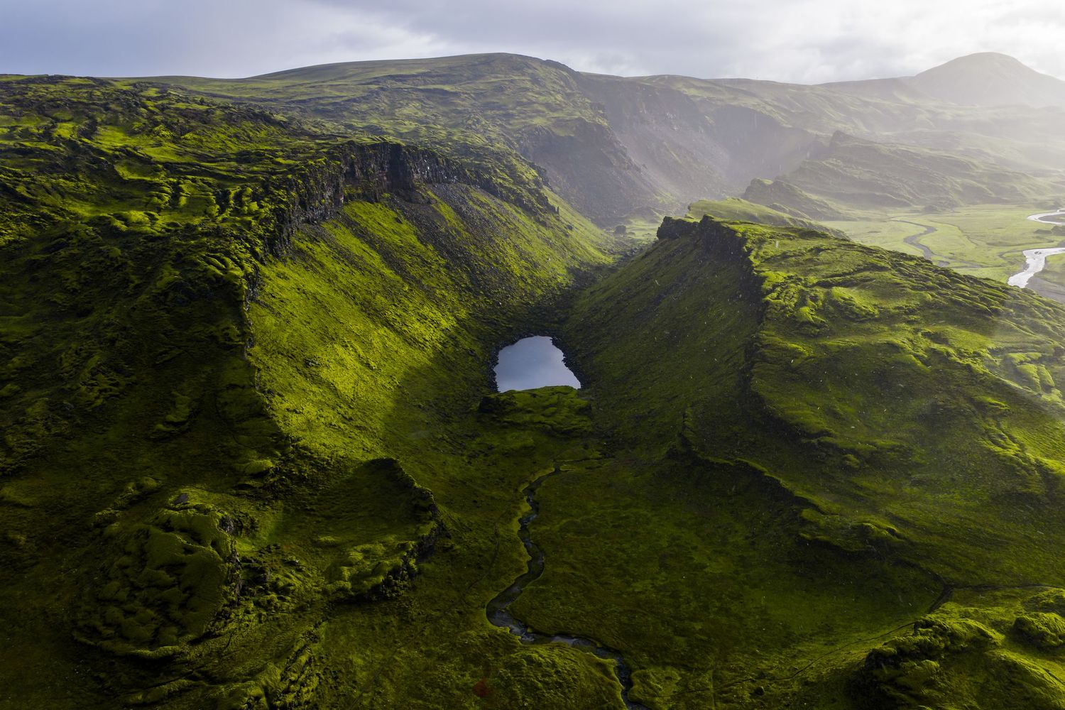 Aerial view of a dramatic natural arch formation in green rolling hills of Iceland's rugged landscape under moody skies.