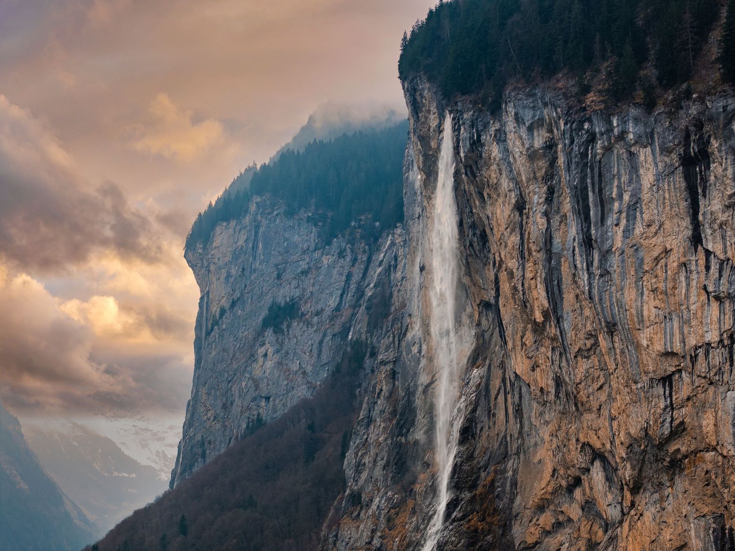 Majestic waterfall streams down massive granite cliff face at sunset with dramatic clouds.