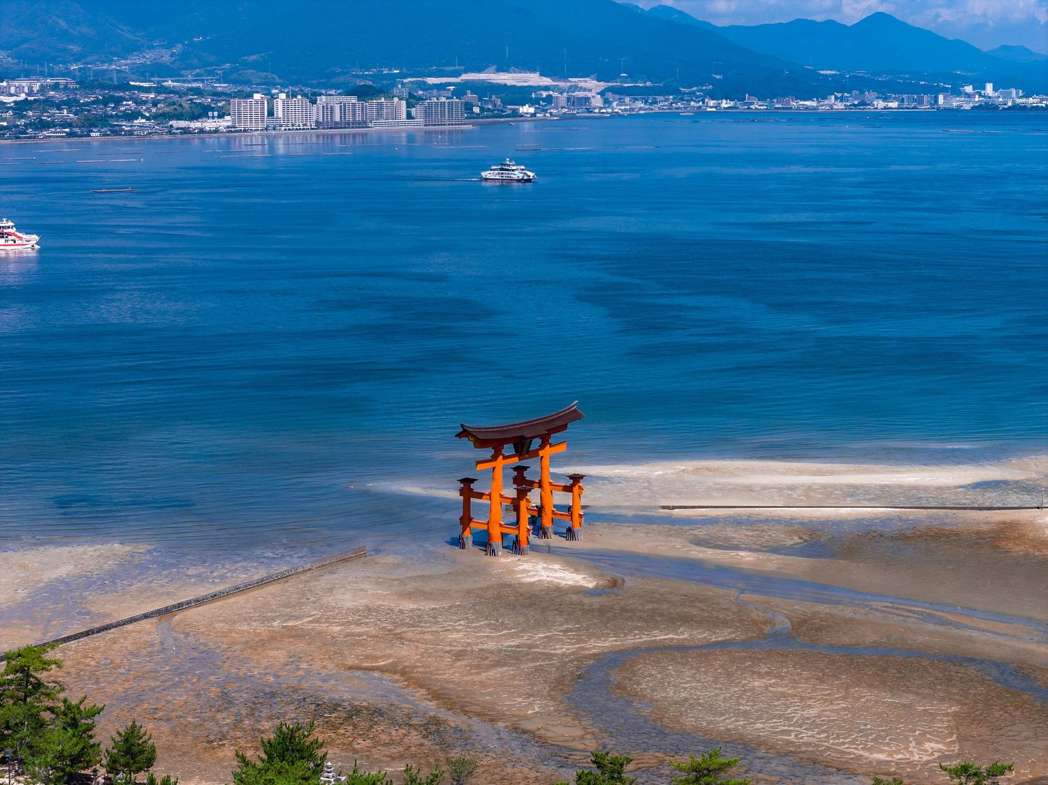 Aerial view of Miyajima's iconic orange torii gate standing in the blue waters off the coast of Japan during low tide.