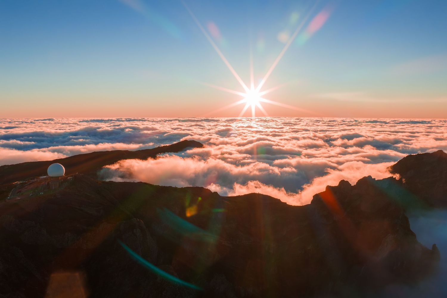 Stunning sunset view above a sea of clouds from mountain summit with sunburst and lens flare effect in golden hour.