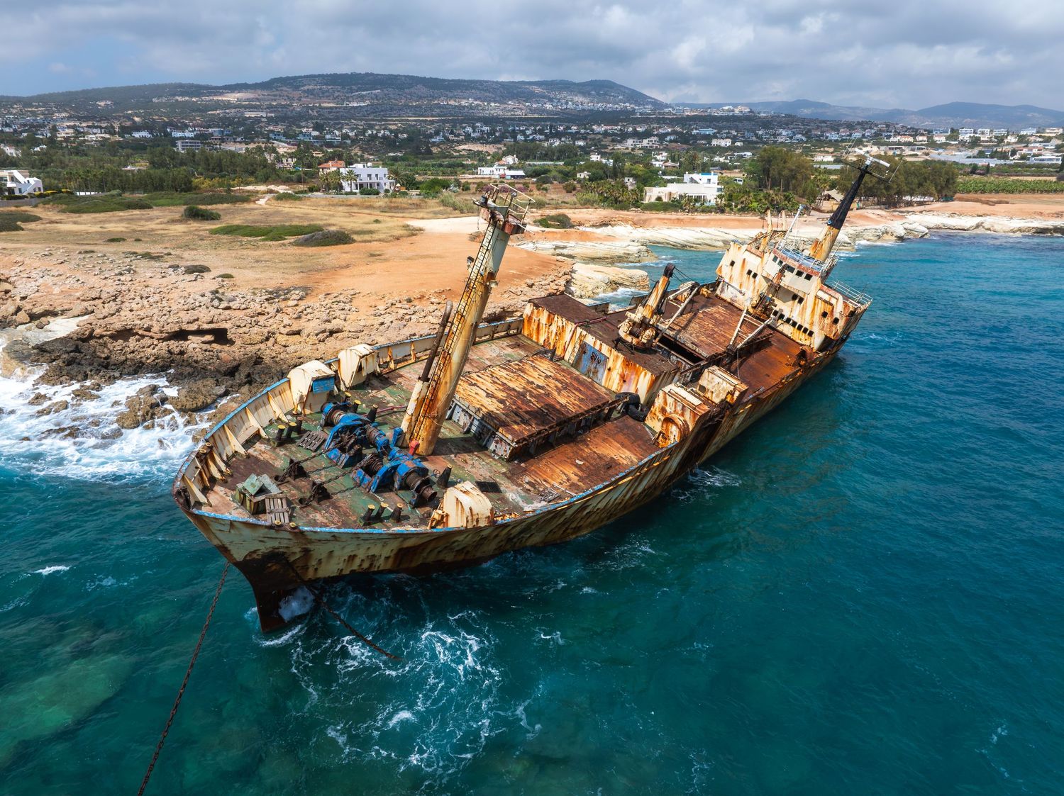 Aerial view of shipwreck on rocky coastline with waves crashing against rusted hull.