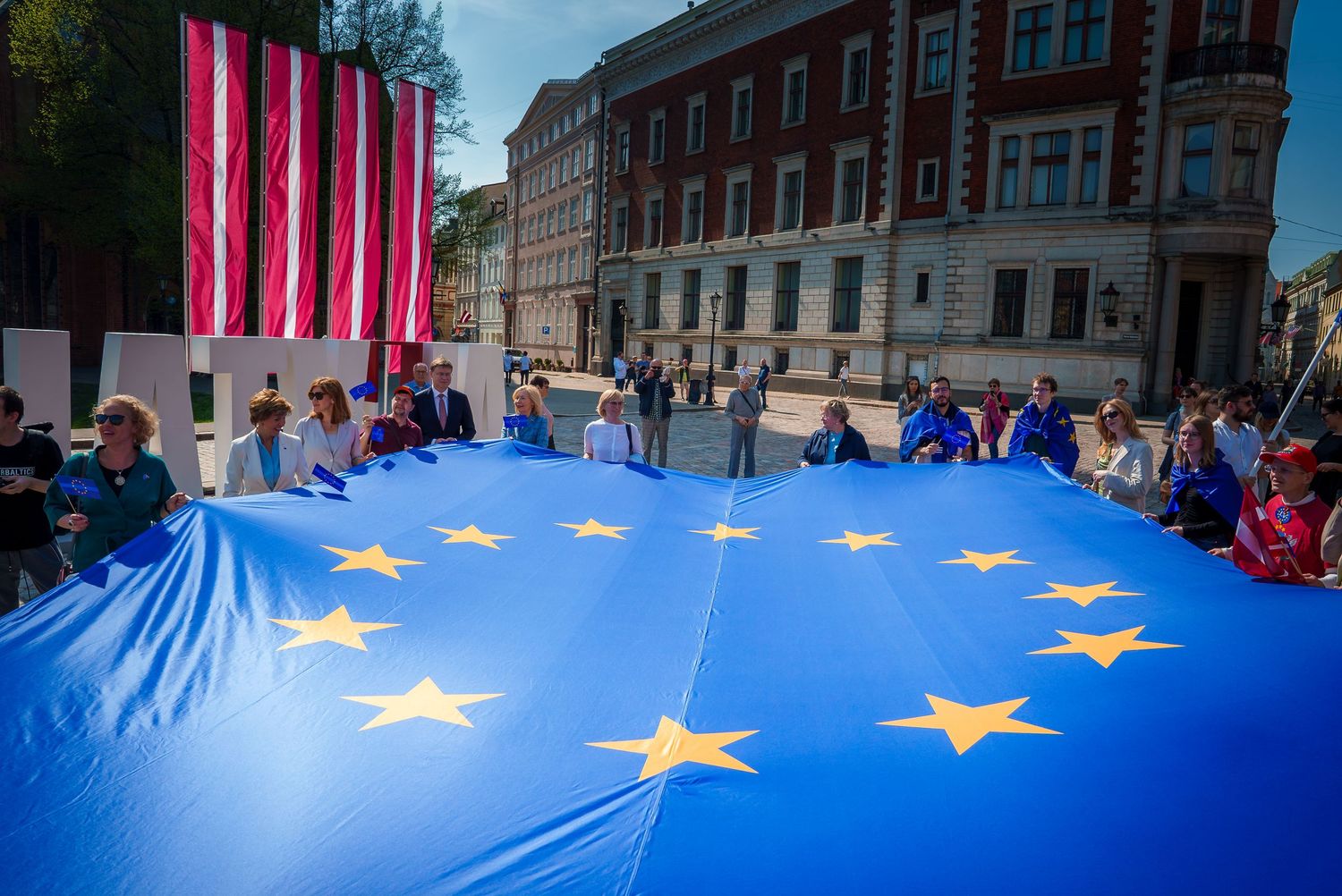 Large European Union flag being held outdoors by multiple people against an urban backdrop.