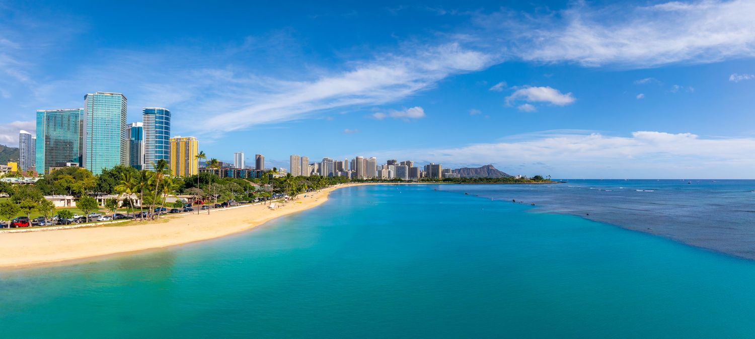 Aerial view of Honolulu's coastal skyline with turquoise waters and sandy beaches in the foreground.