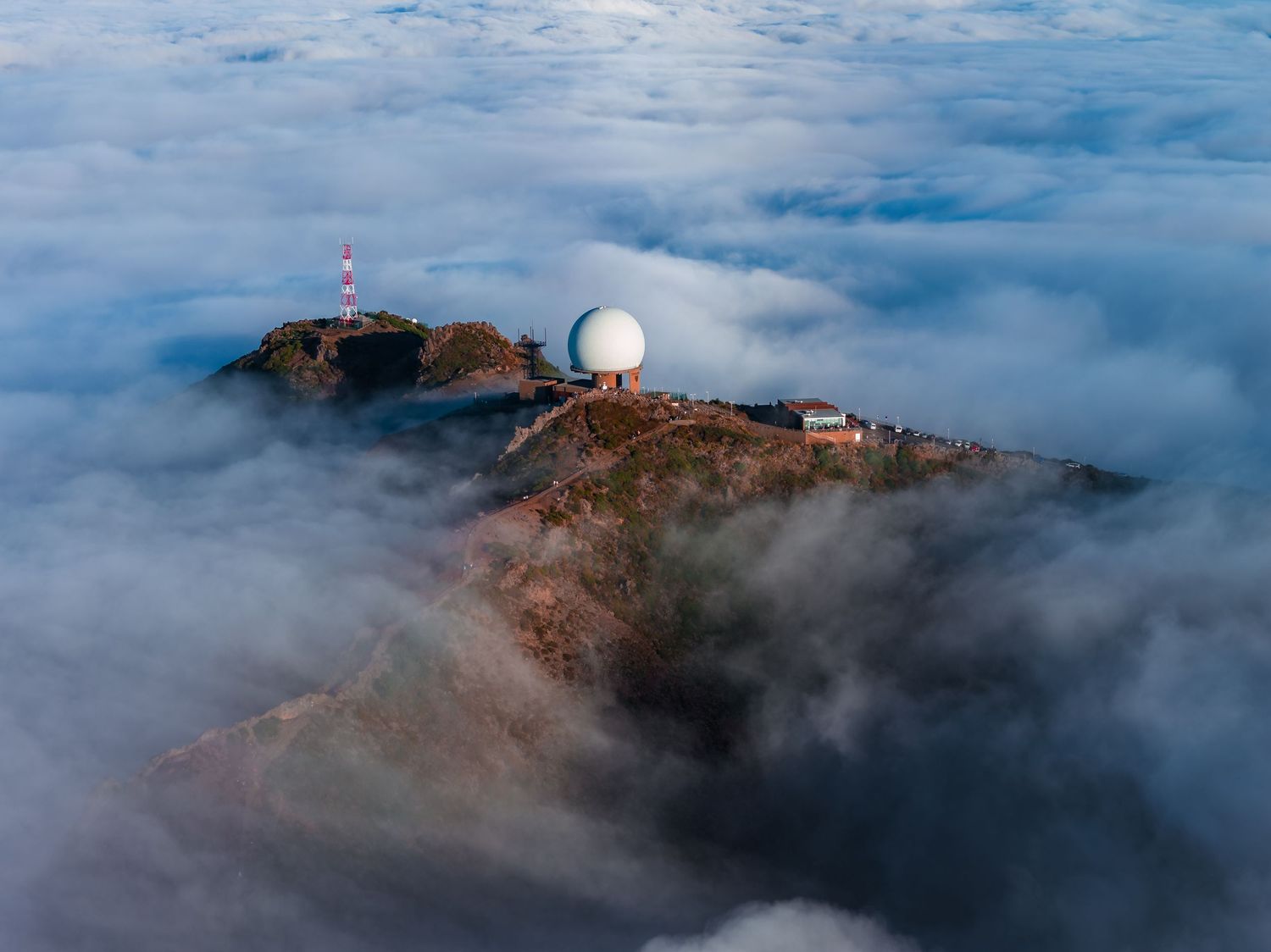 Weather radar station on mountain peak rises above dramatic sea of clouds during foggy conditions.