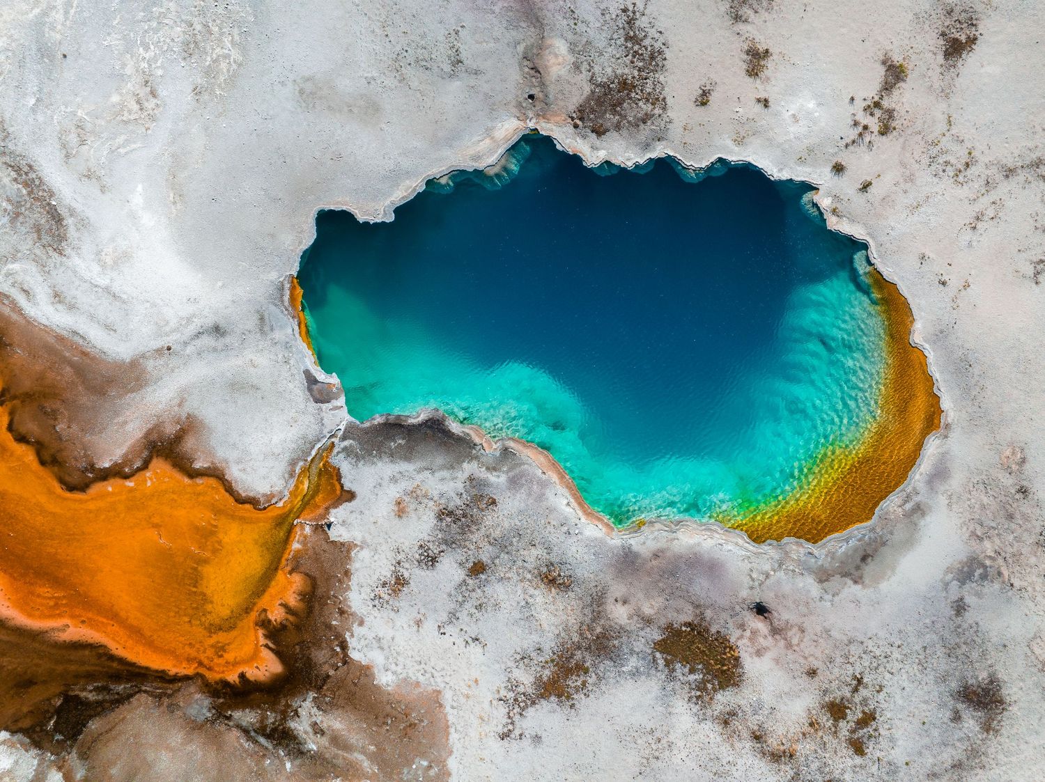 Aerial photograph of a turquoise hot spring with orange mineral deposits in a desert landscape.
