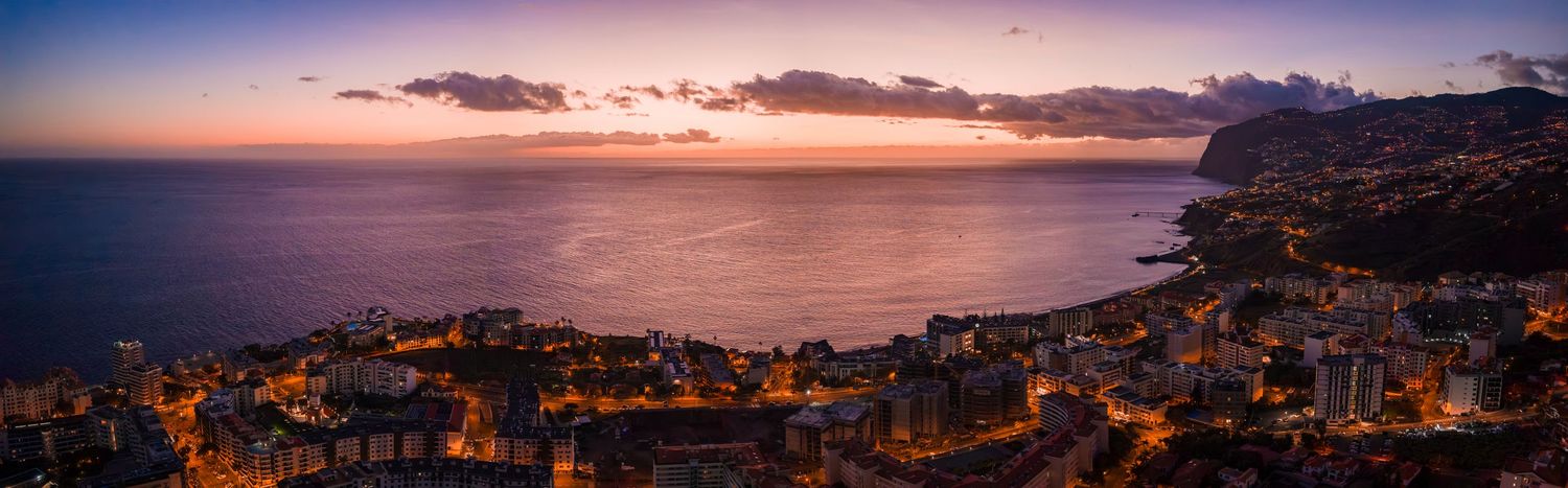Panoramic sunset view over a coastal Mediterranean town with glowing orange sky and rugged hillside buildings.