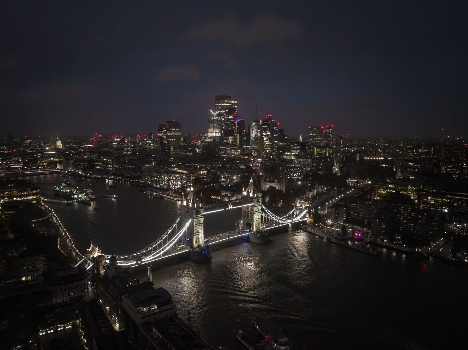Aerial panorama of London at night showing illuminated Tower Bridge and city skyline along the River Thames.