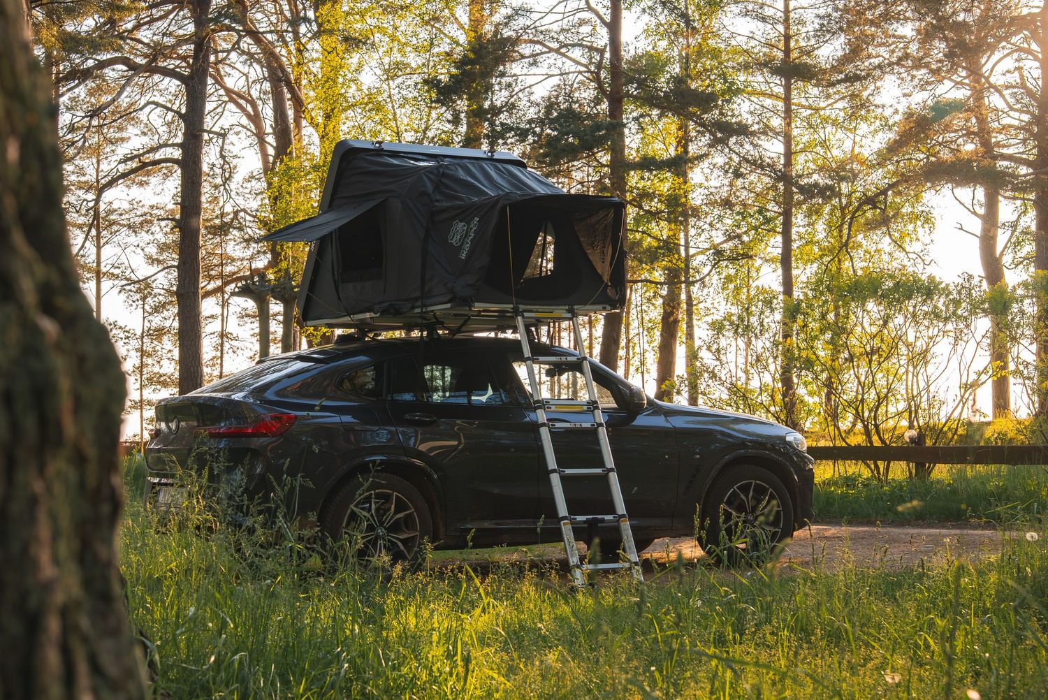 Black vehicle with rooftop tent parked in forest clearing at sunset.