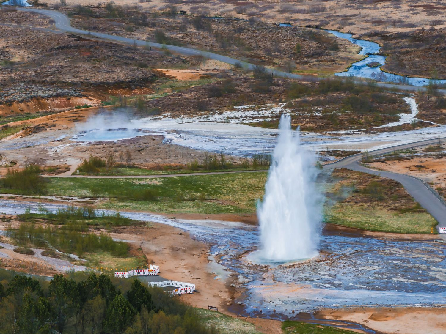Geothermal geyser erupting with steam and hot water in a rocky landscape with winding rivers.