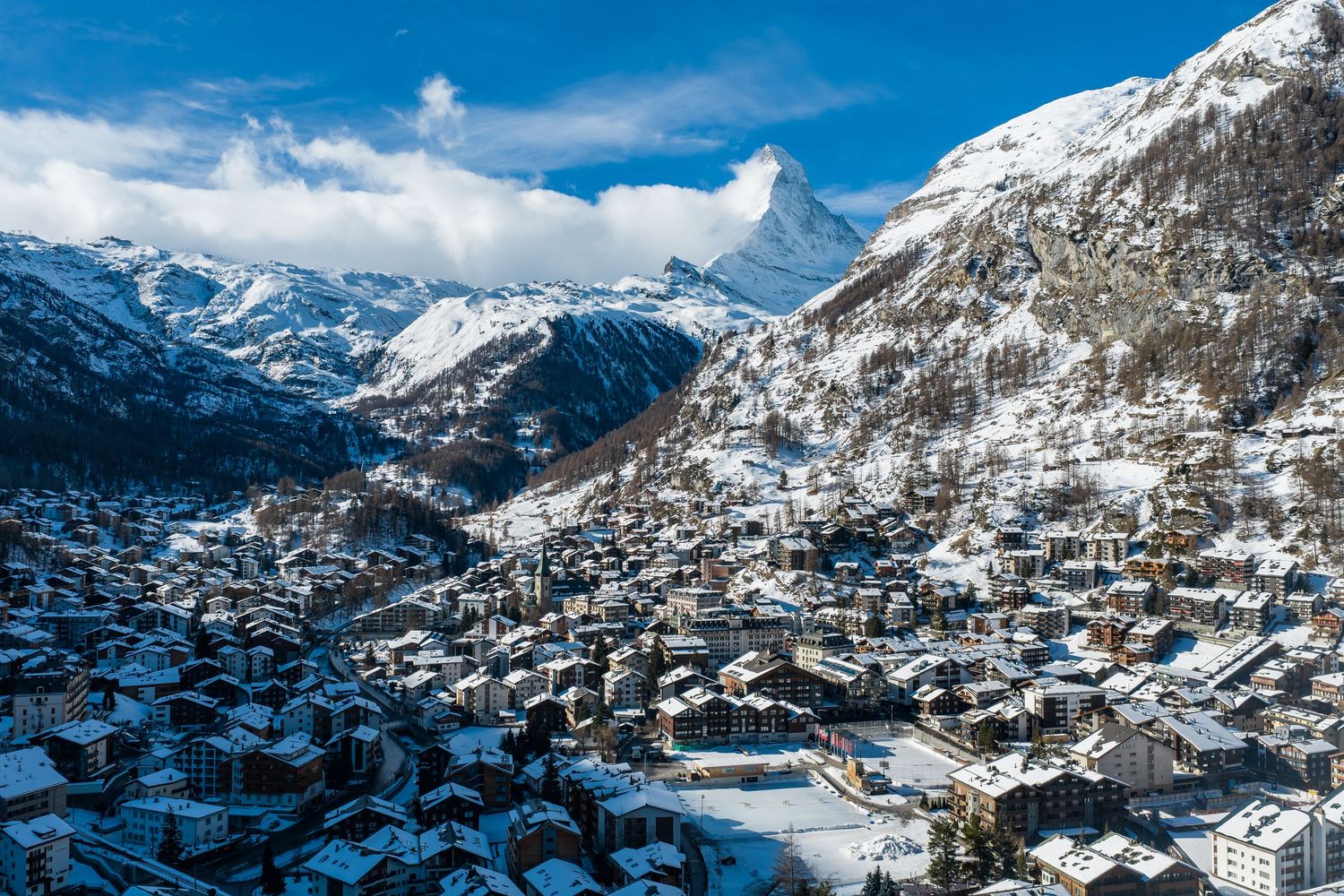 Aerial panoramic view of a snow-covered alpine village nestled in the mountains with chalets and ski slopes.