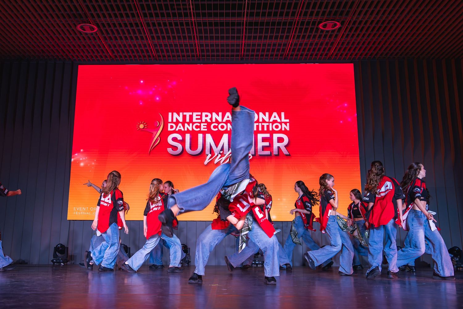 Dance performance on stage with dramatic red lighting and white-clad performers in formation.