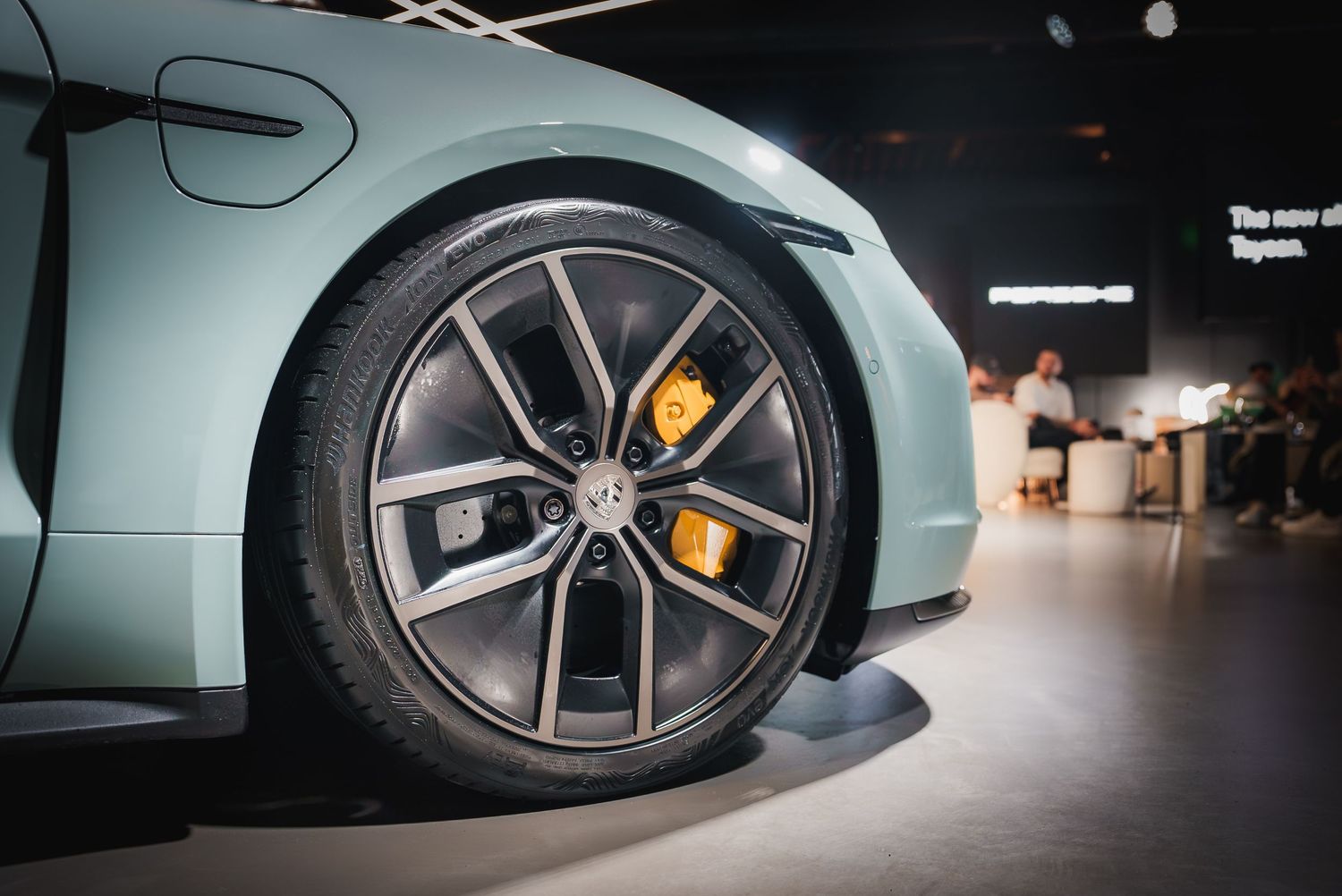Detail shot of a Porsche Taycan's wheel and brake caliper in a showroom setting.