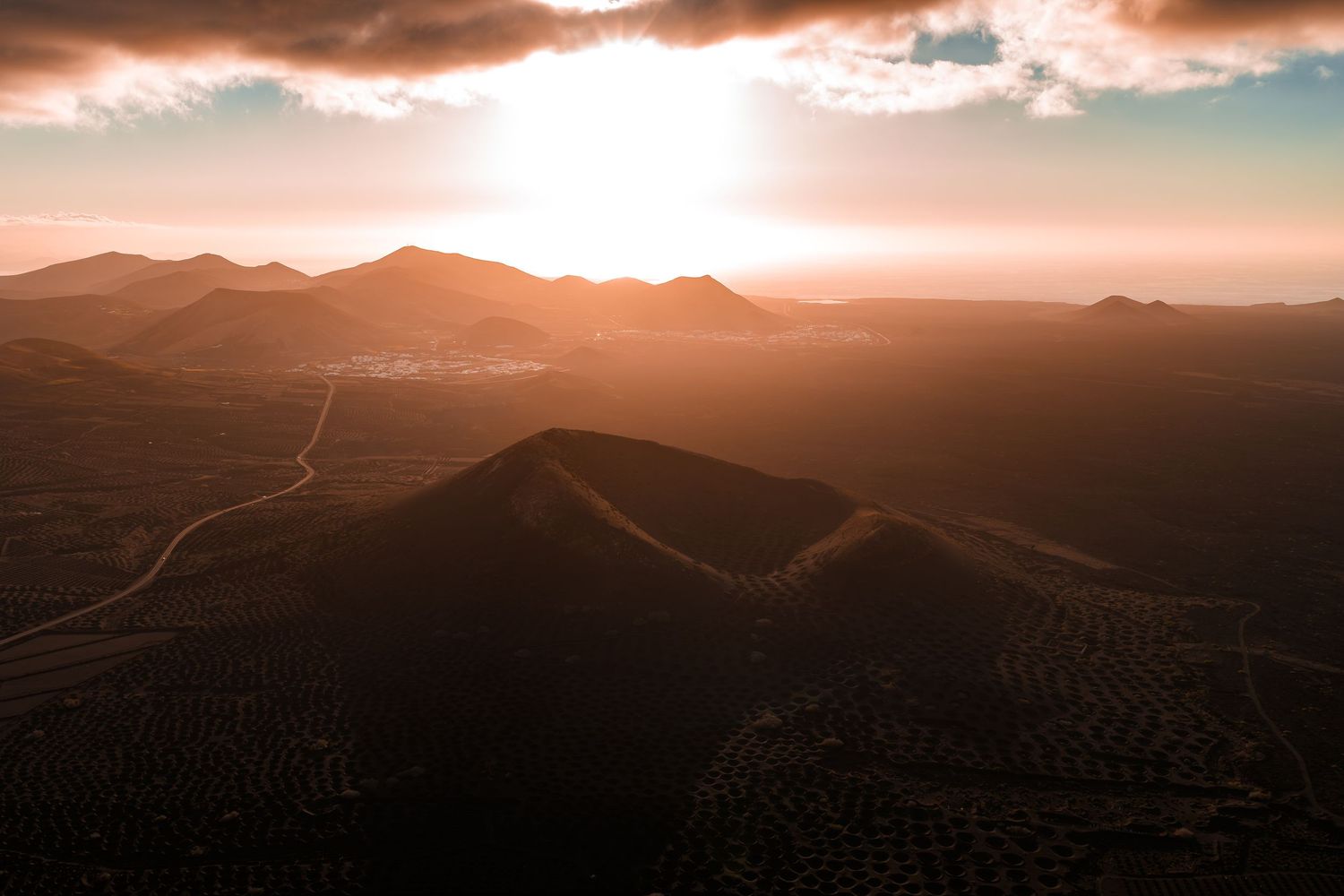 Dramatic sunset over mountain ranges with winding road through desert landscape in golden hour light.