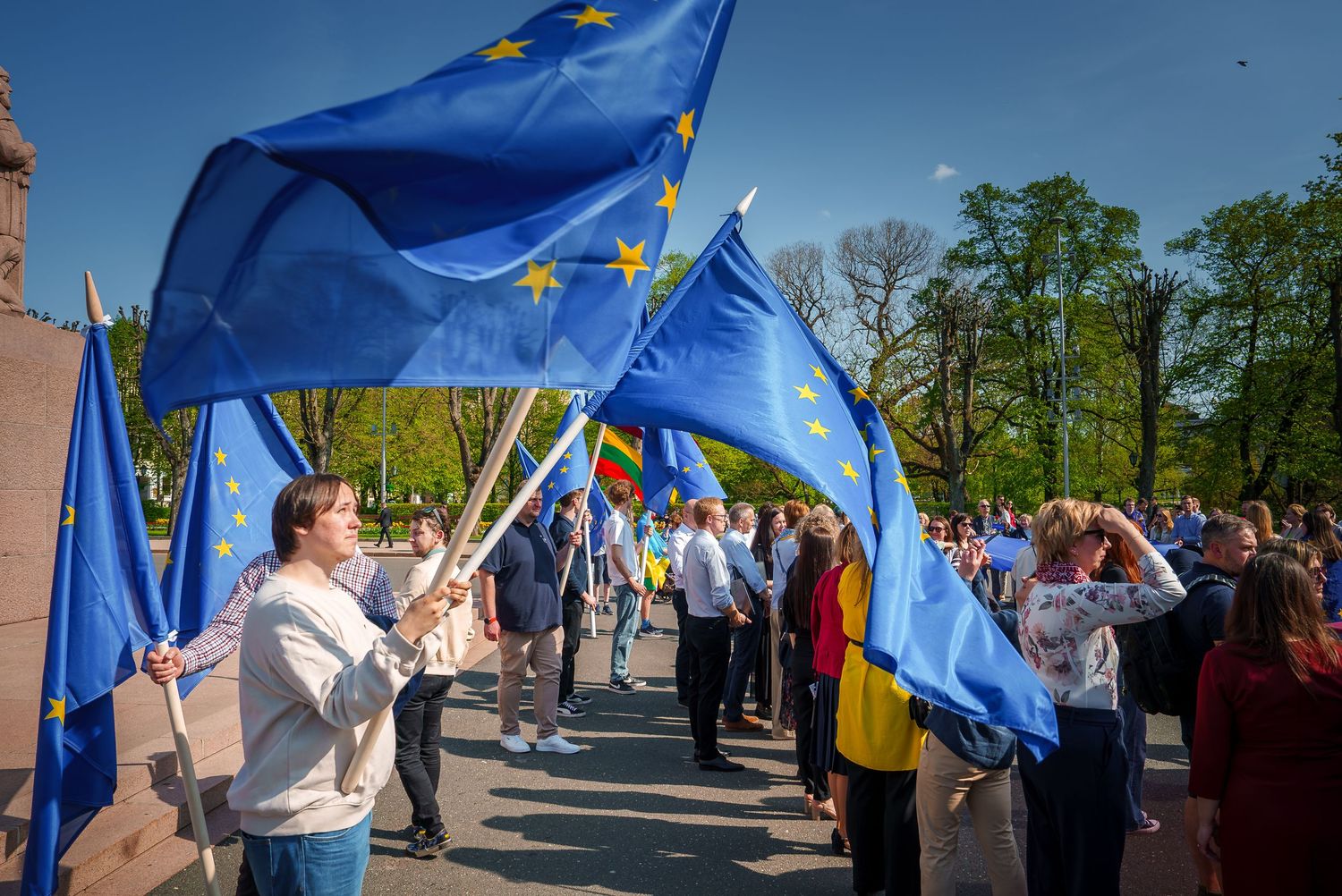 Crowd waving European Union flags during outdoor gathering on sunny day.
