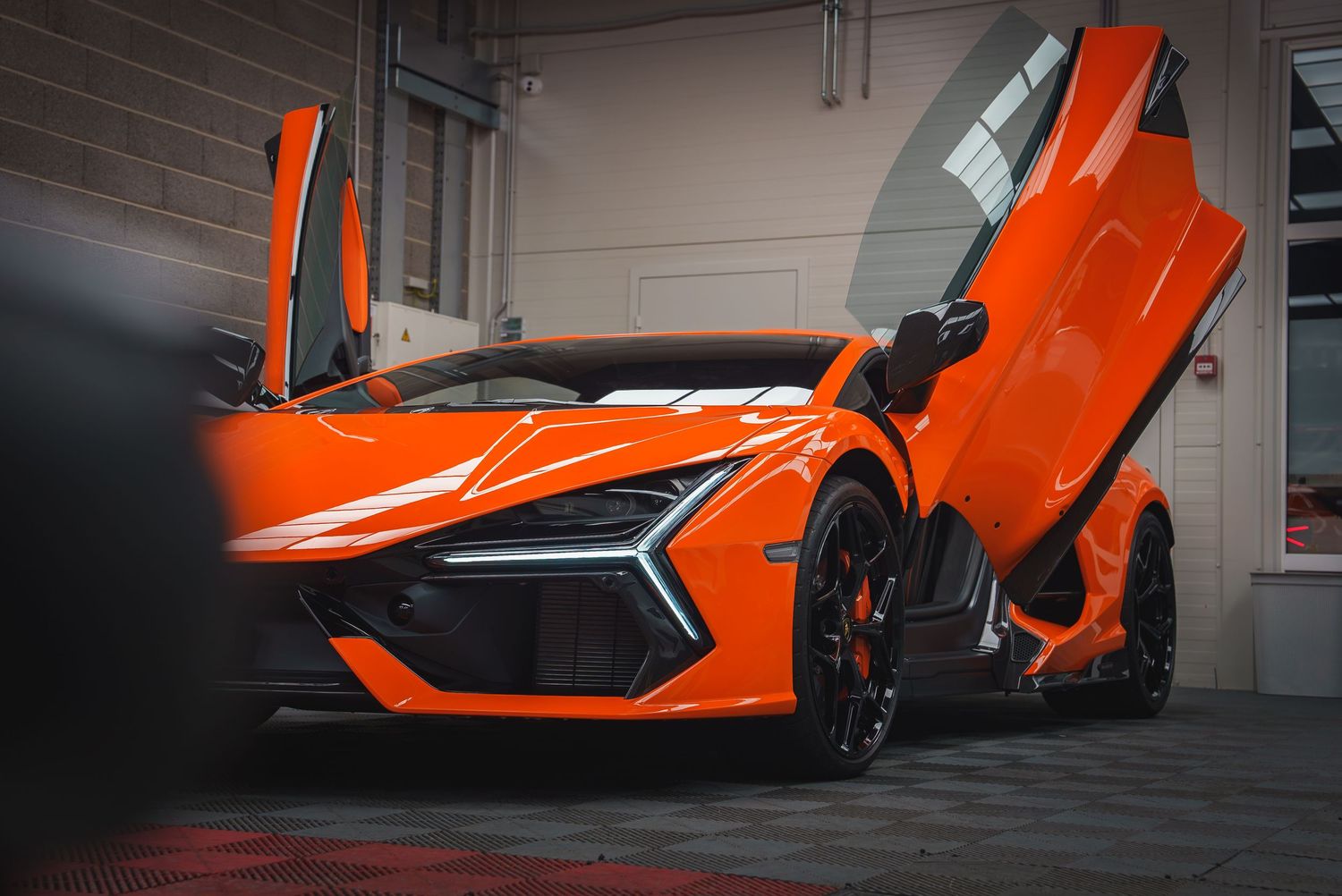 Bright orange Lamborghini supercar with scissor doors displayed in a minimalist garage setting.