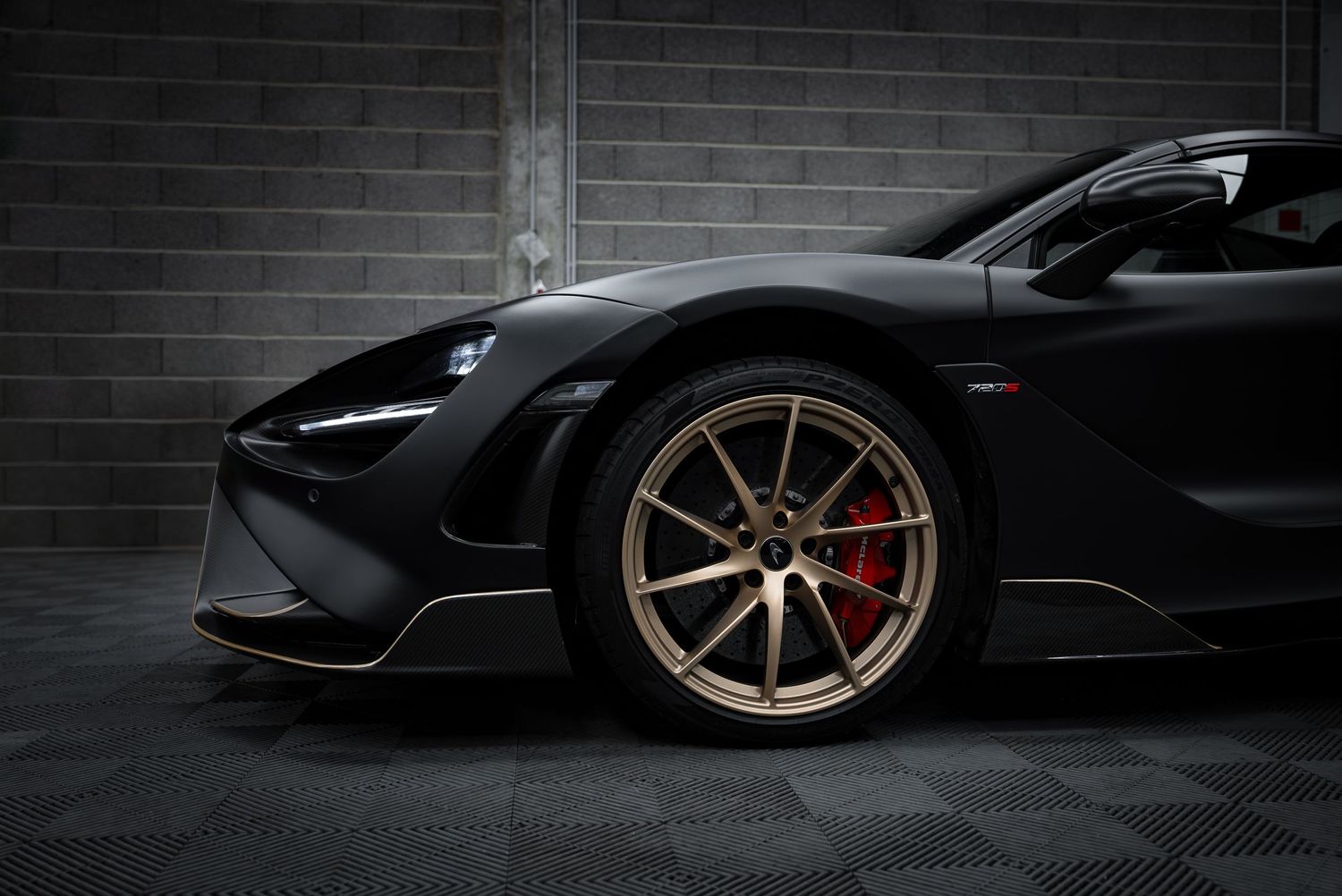 Black McLaren sports car with bronze wheels and red brake calipers against dark brick wall.