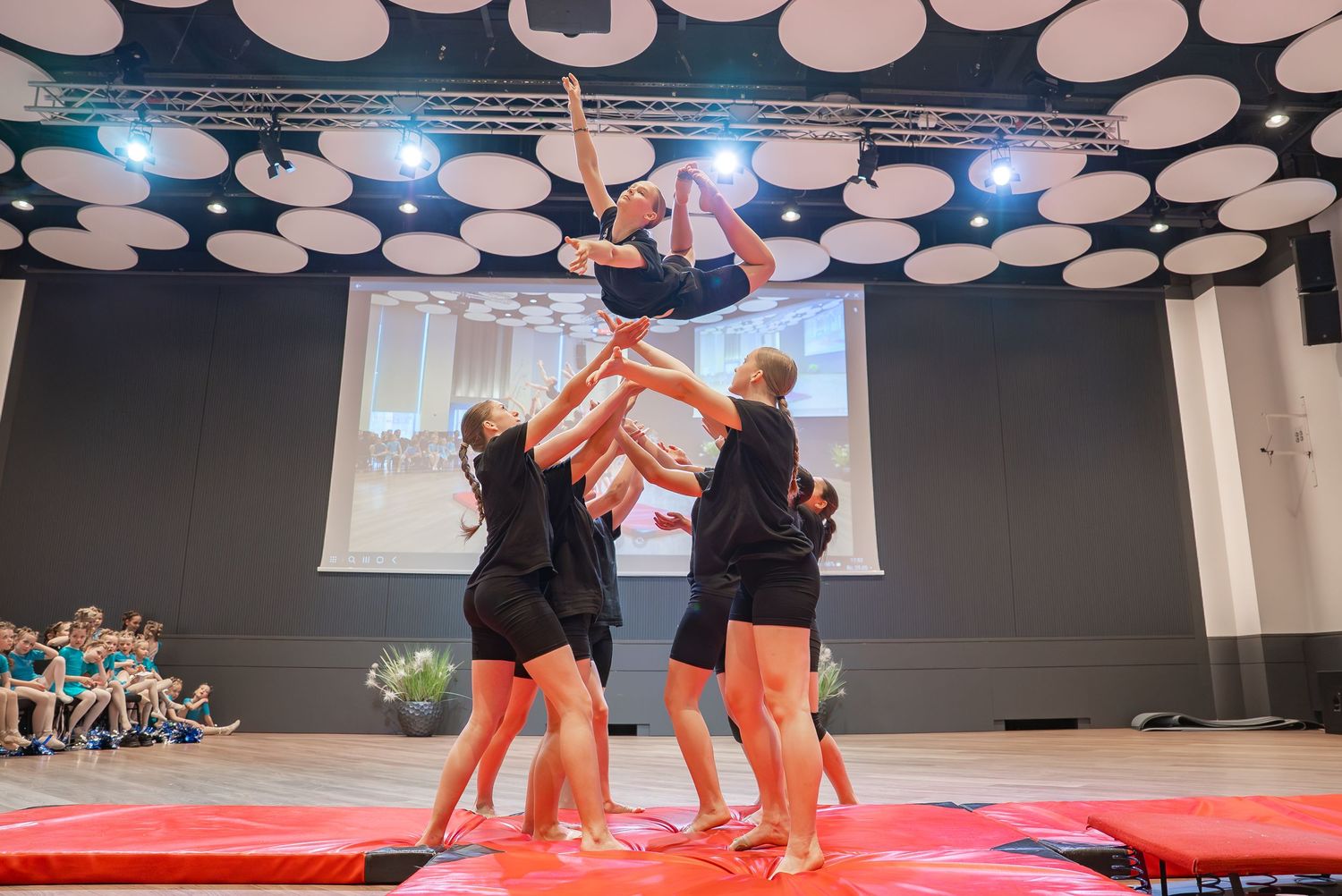Group performing an acrobatic lift routine on a red mat under circular ceiling lights.