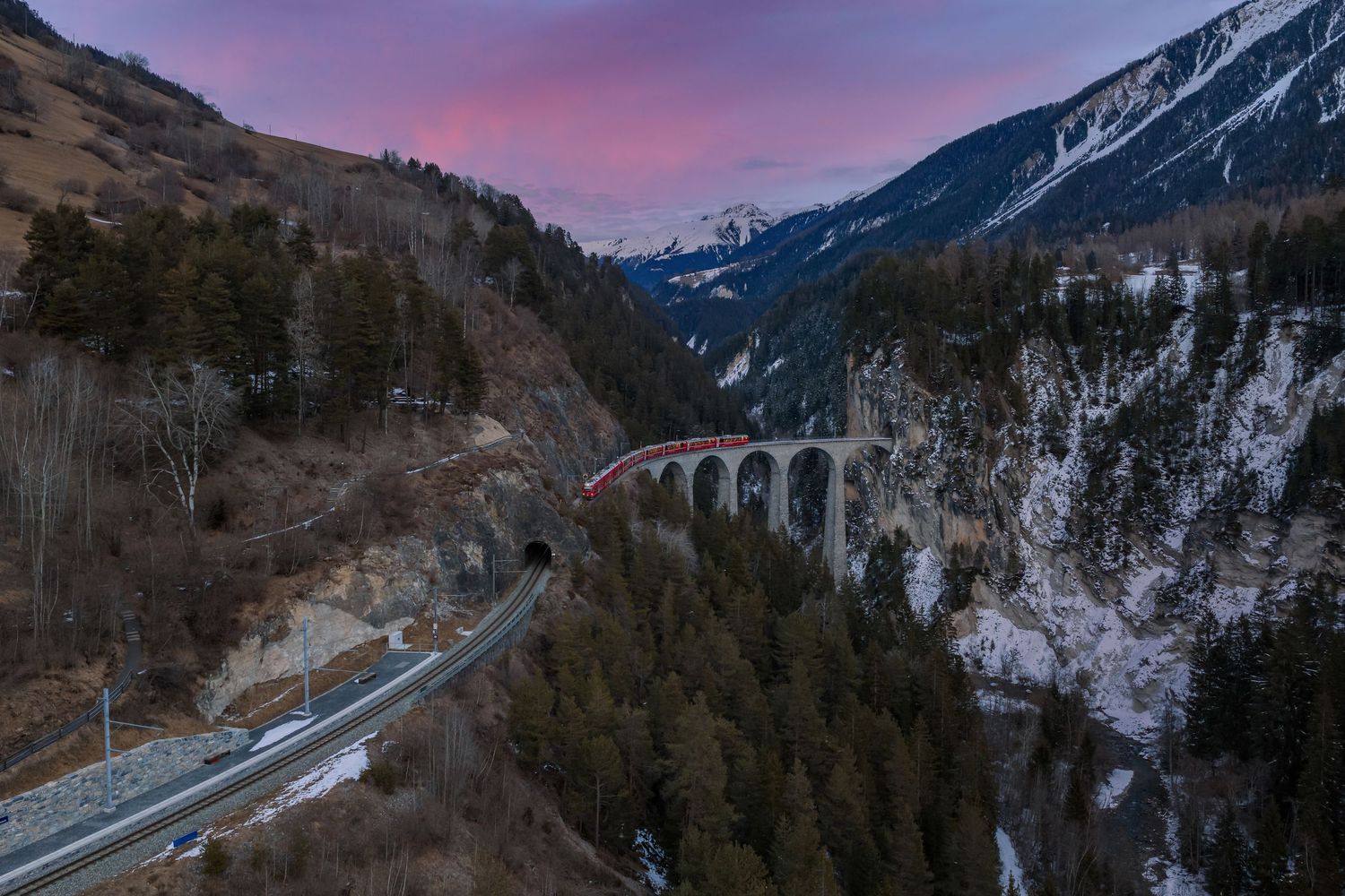 Snowy mountain road curves through a dramatic winter landscape at sunset with purple and pink sky.