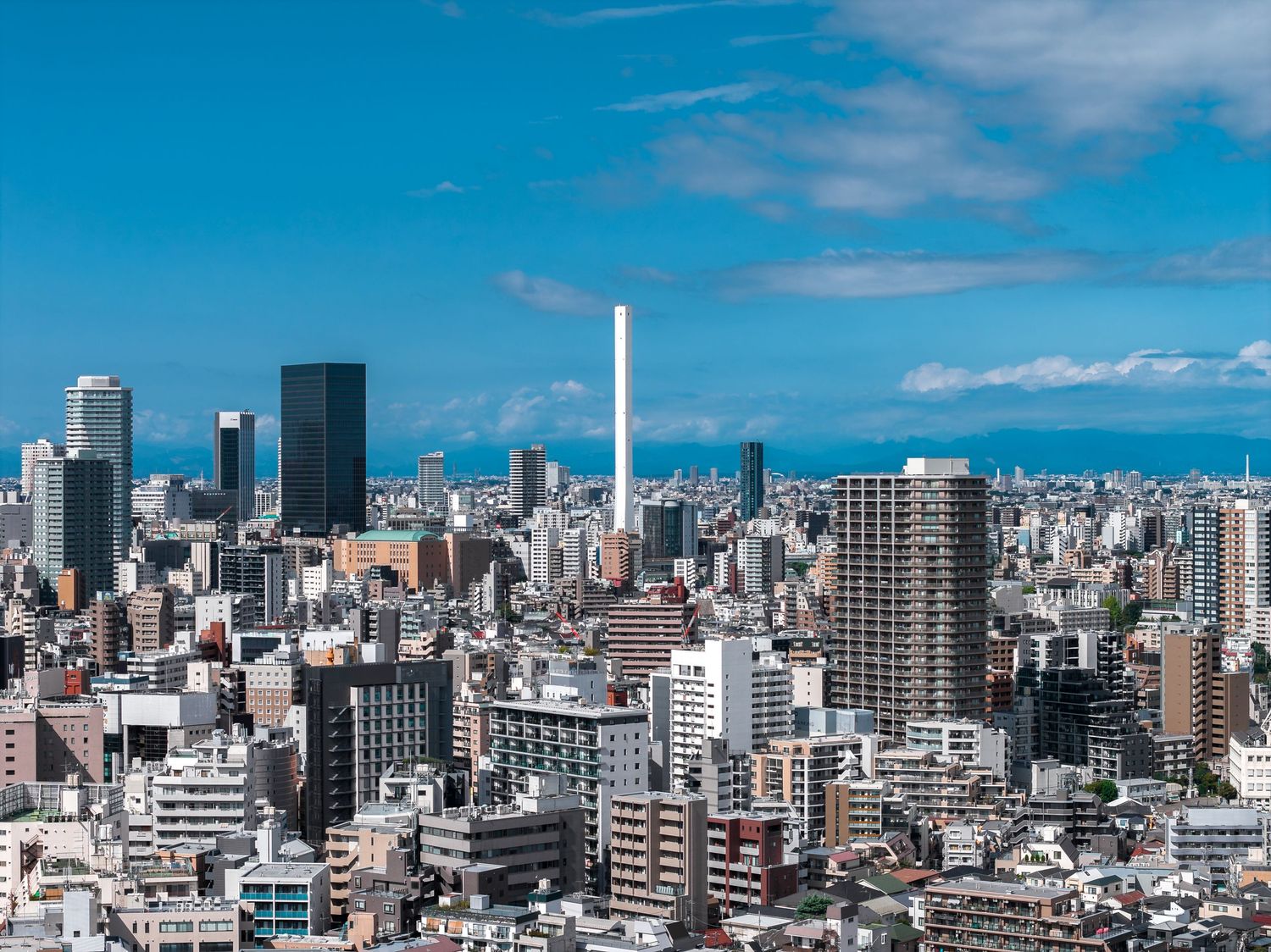 A panoramic cityscape view of a modern Japanese metropolis with tall buildings against a bright blue sky.