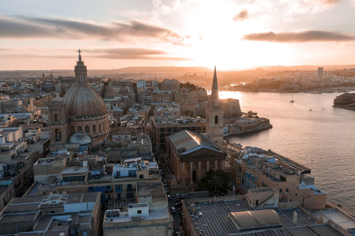 Panoramic sunset view of Valletta, Malta's historic skyline with domed churches and Mediterranean Sea in golden light.