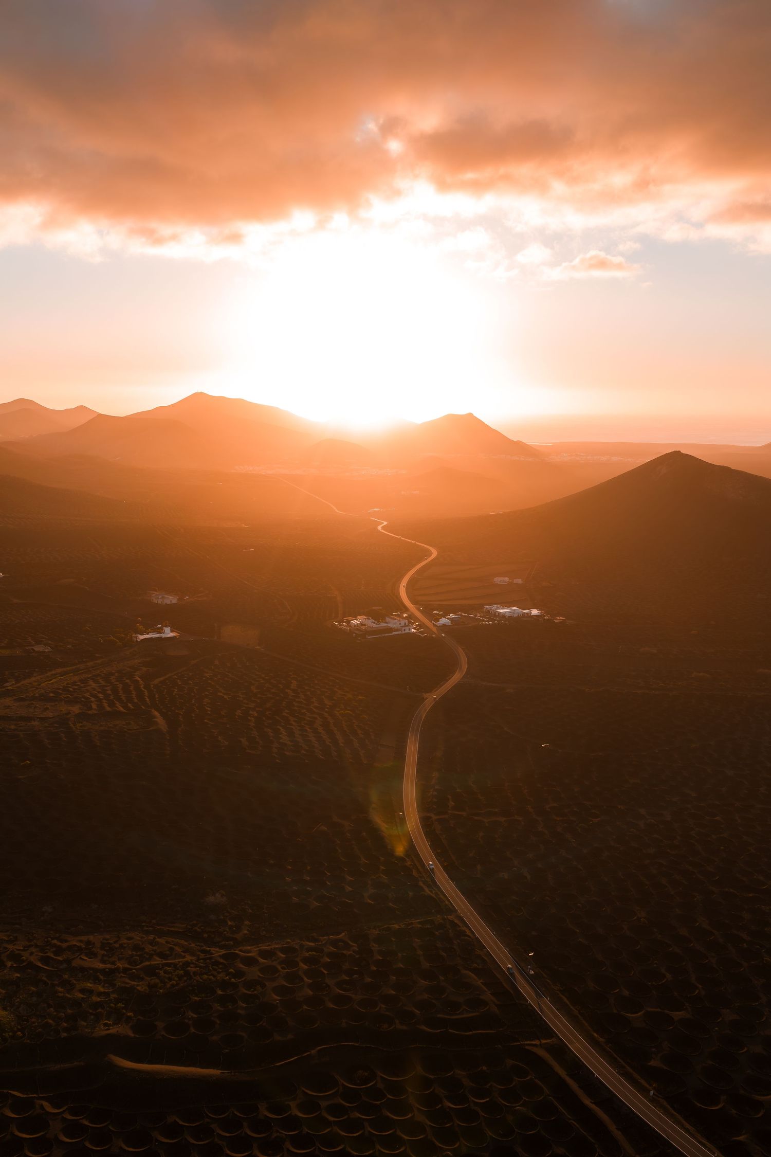 Winding mountain road cuts through rugged terrain at sunset with dramatic orange sky and mountain silhouettes.