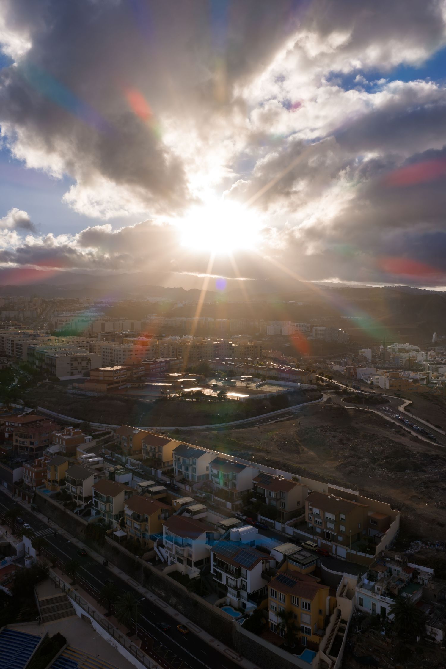 Dramatic aerial panorama of a cityscape at sunset with sunbeams breaking through stormy clouds over urban buildings.