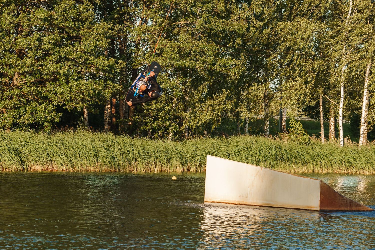 Lake wakeboarding park with ramps and obstacles surrounded by green trees.