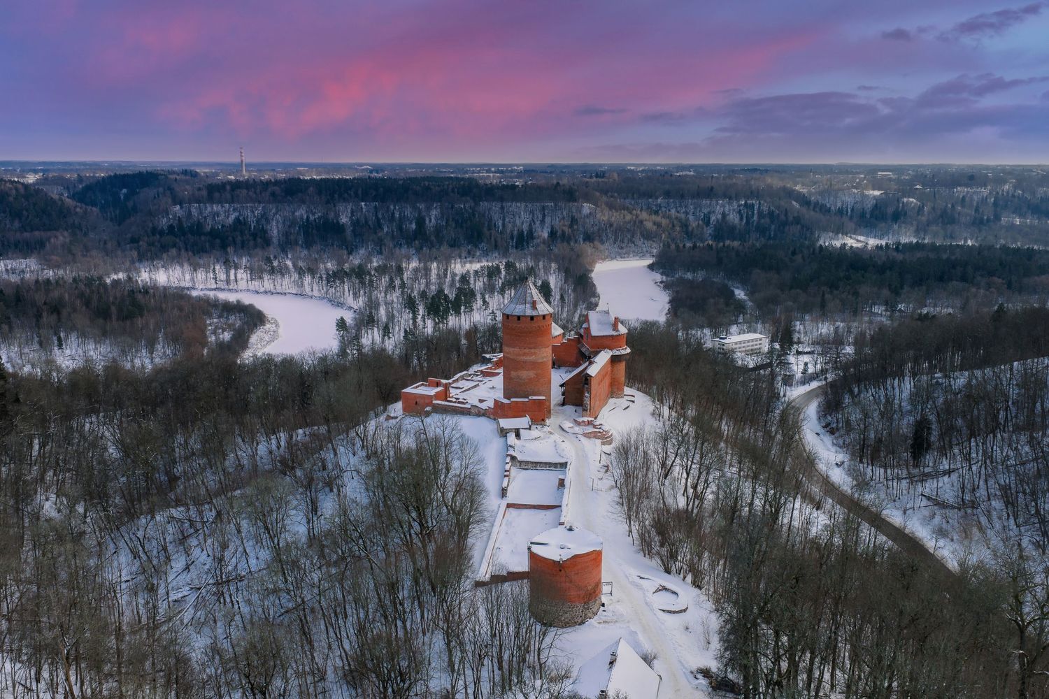 Aerial view of snowy winter landscape with buildings and trees under a vibrant pink and purple sunset sky.
