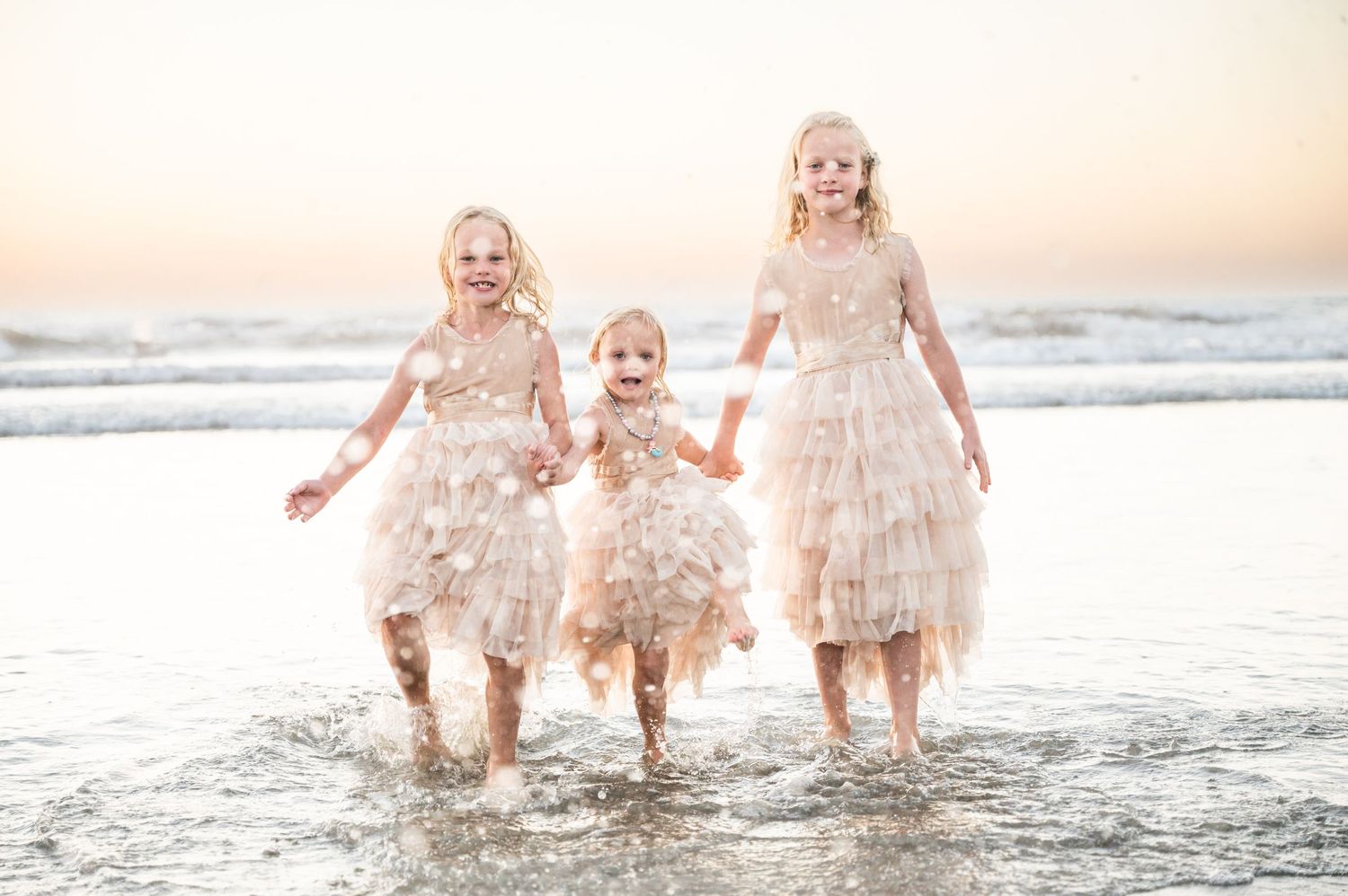 Three girls in soft pink dresses playing in the ocean, kicking up water.