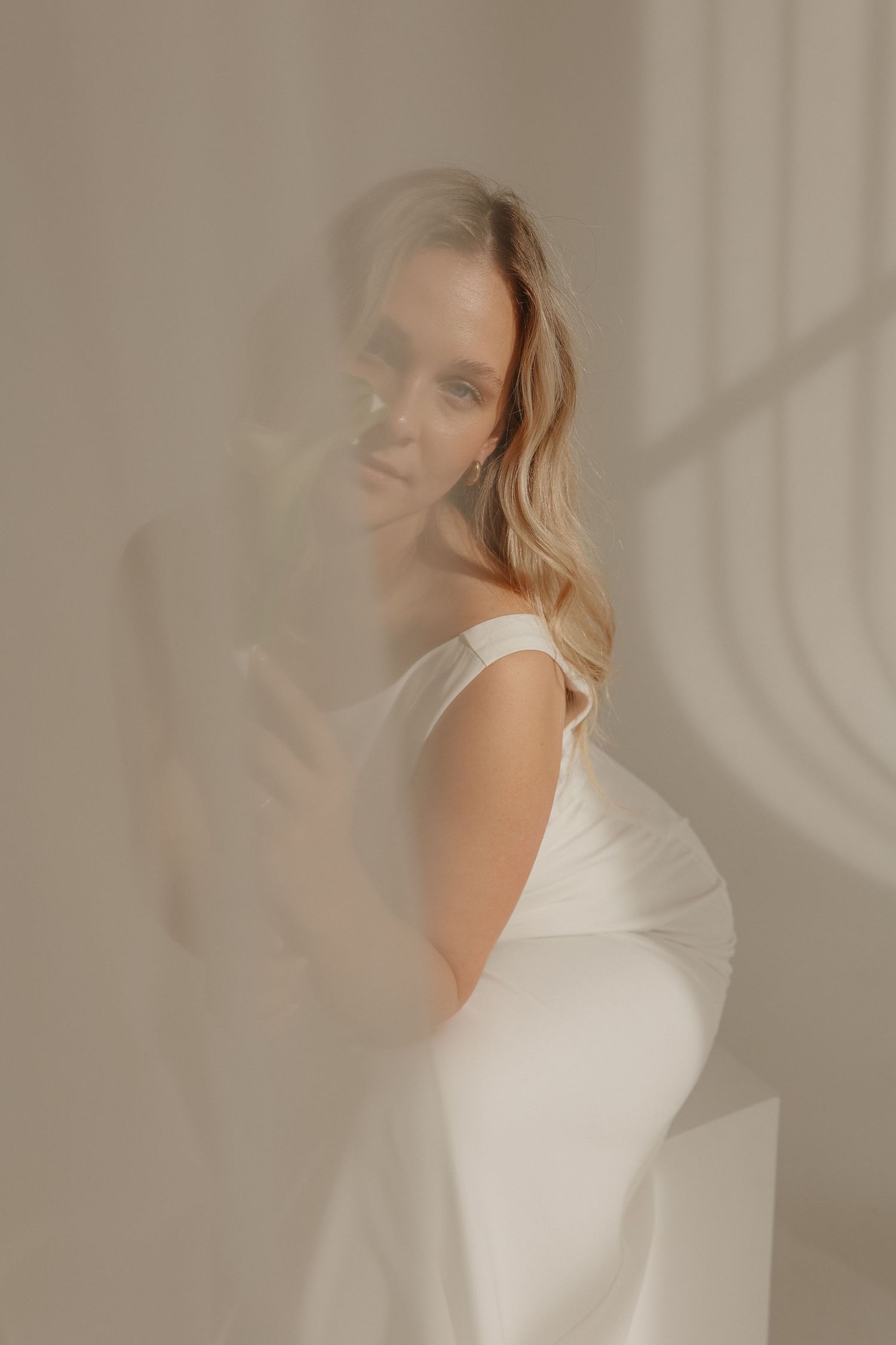Bride in minimalist white off-shoulder satin gown photographed through sheer fabric in soft light.
